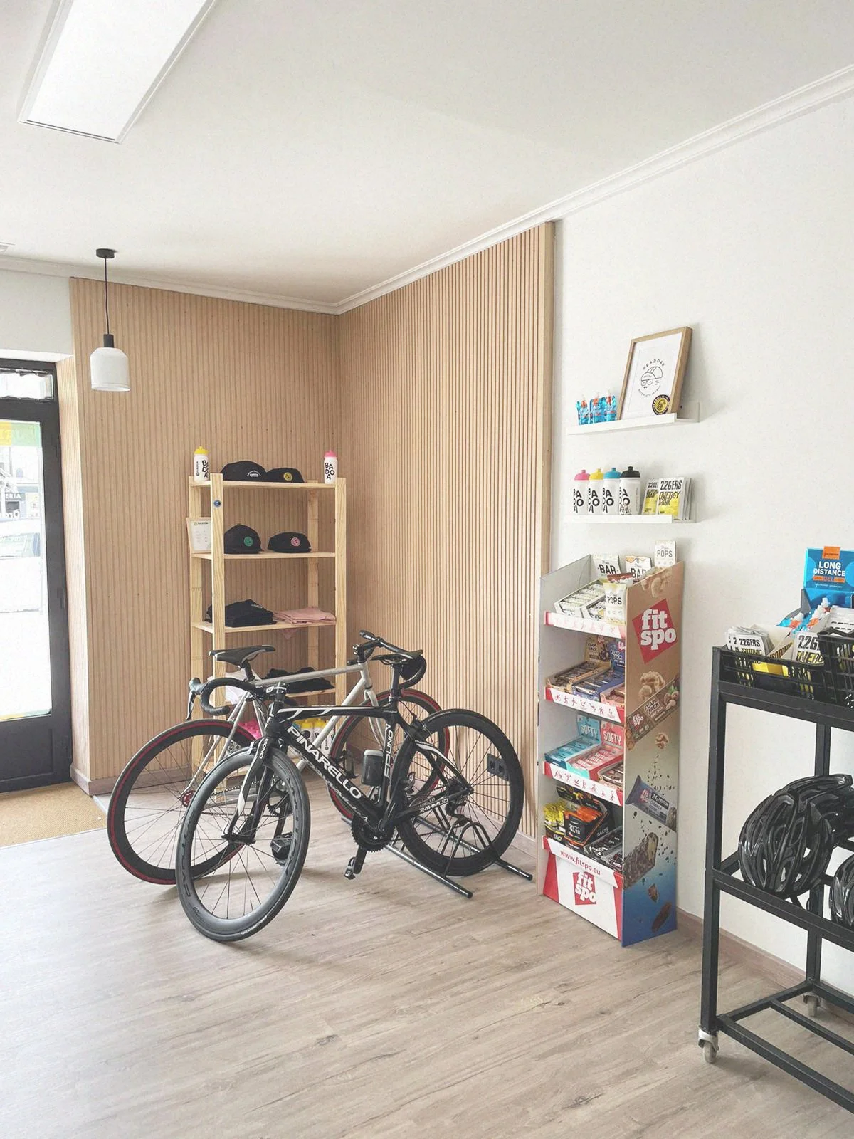 Interior de una tienda con dos bicicletas de ruta, estante con gorras, una estantería de snacks y bebidas de proteína, y una exhibición de cascos en una estantería de metal.