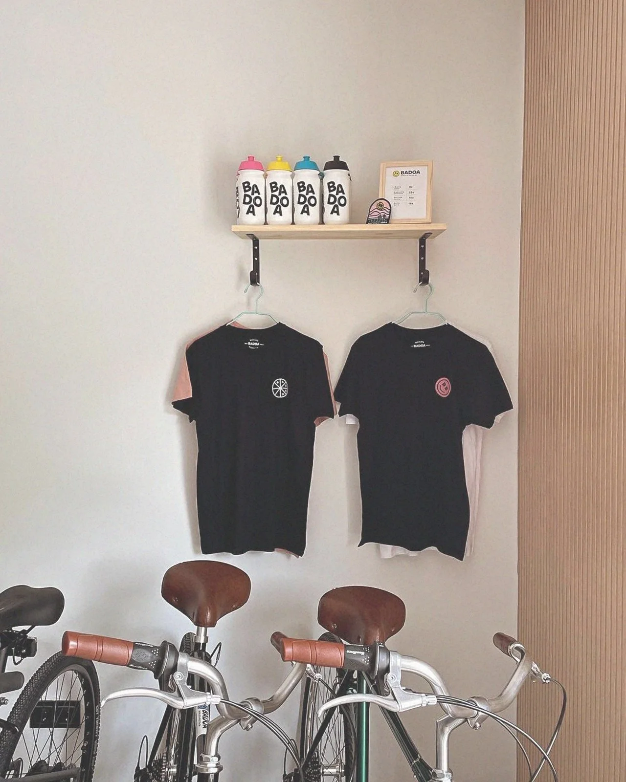Cuatro botellas de agua con etiquetas que dicen 'BADOA' en la parte superior. Dos camisetas negras con logotipos en el centro colgadas en colgadores y bicicletas estacionadas en primer plano.