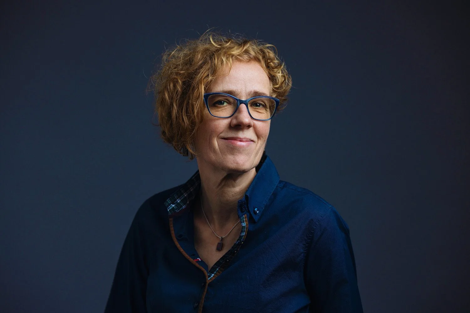 Ein Porträt einer lächelnden Frau mit kurzen, lockigen roten Haaren und blauen Brillen, die dunkle Kleidung trägt, vor einem dunklen Hintergrund.