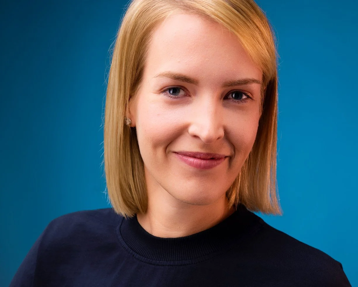 Nahaufnahme einer lächelnden jungen Frau mit blonden Haaren vor einem blauen Hintergrund.