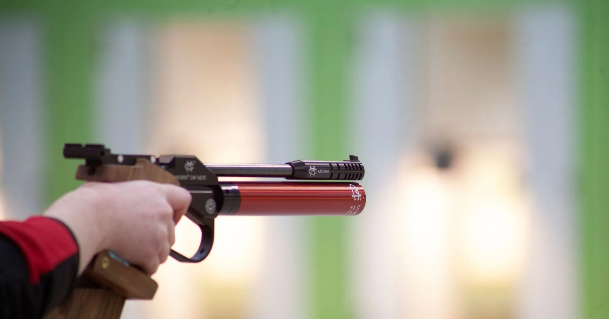 A person aiming an air rifle with a red accent colour at a shooting range, with targets blurred in the background.