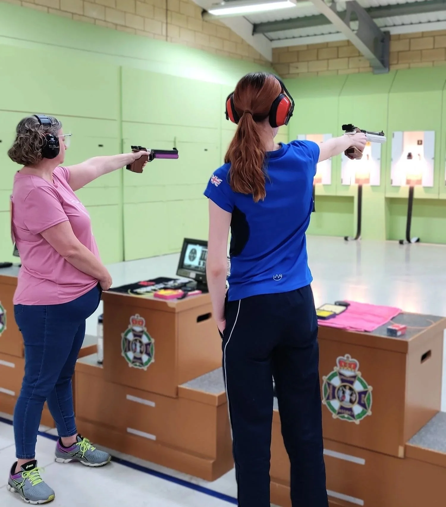 Two women participating in a shooting competition at an indoor range, both aiming handguns at targets, with ear protection