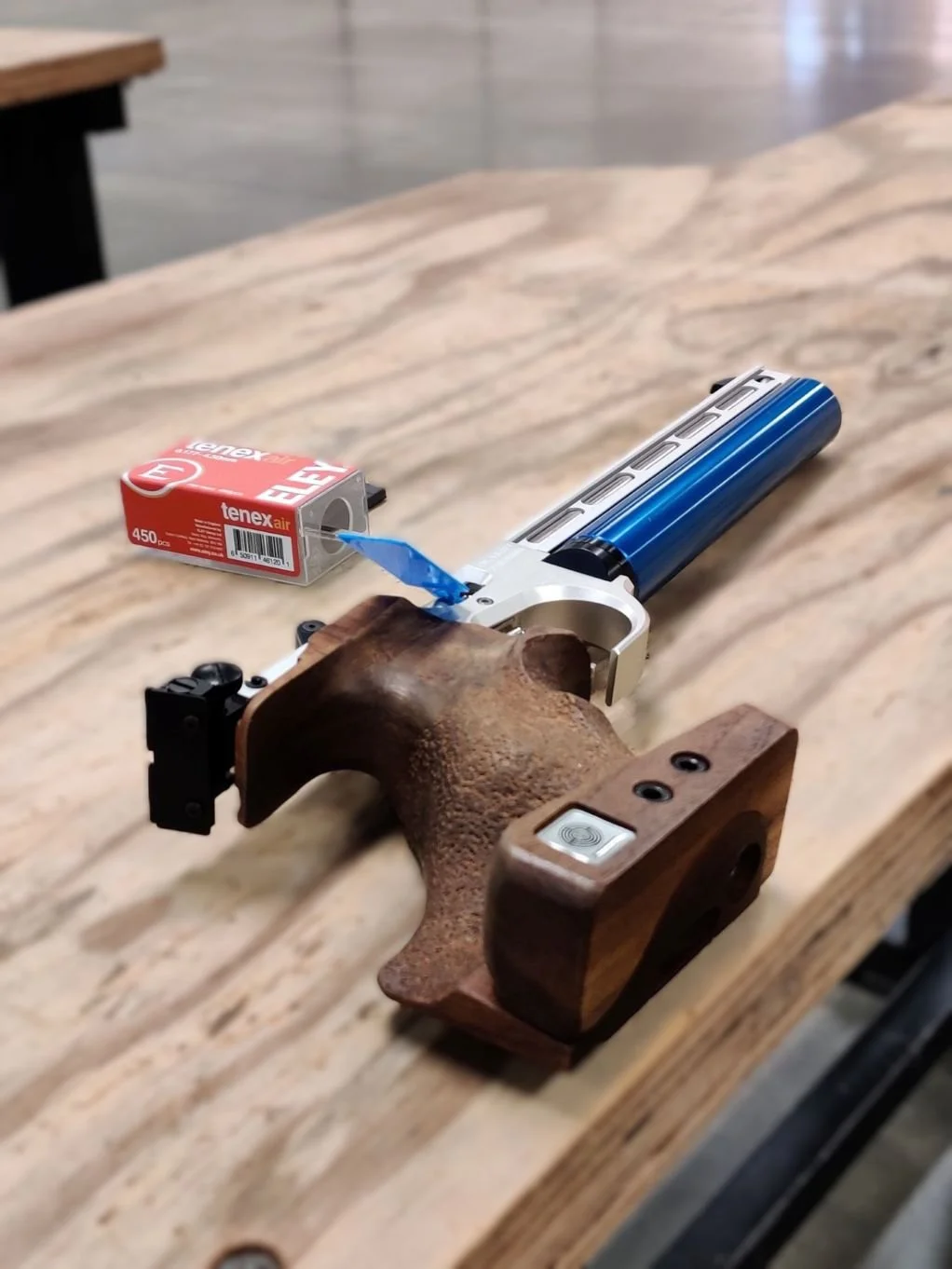 A pistol with a wooden looking grip, metallic slide, and a blue canister attached. There is a box of ELEY TENEX air pellets nearby on a wooden surface.