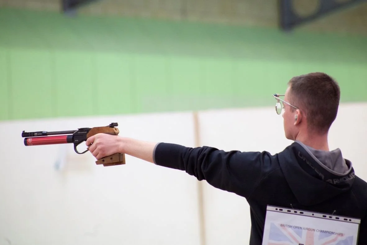 A young man wearing glasses and a black jacket competing in a target shooting event at an indoor shooting range.