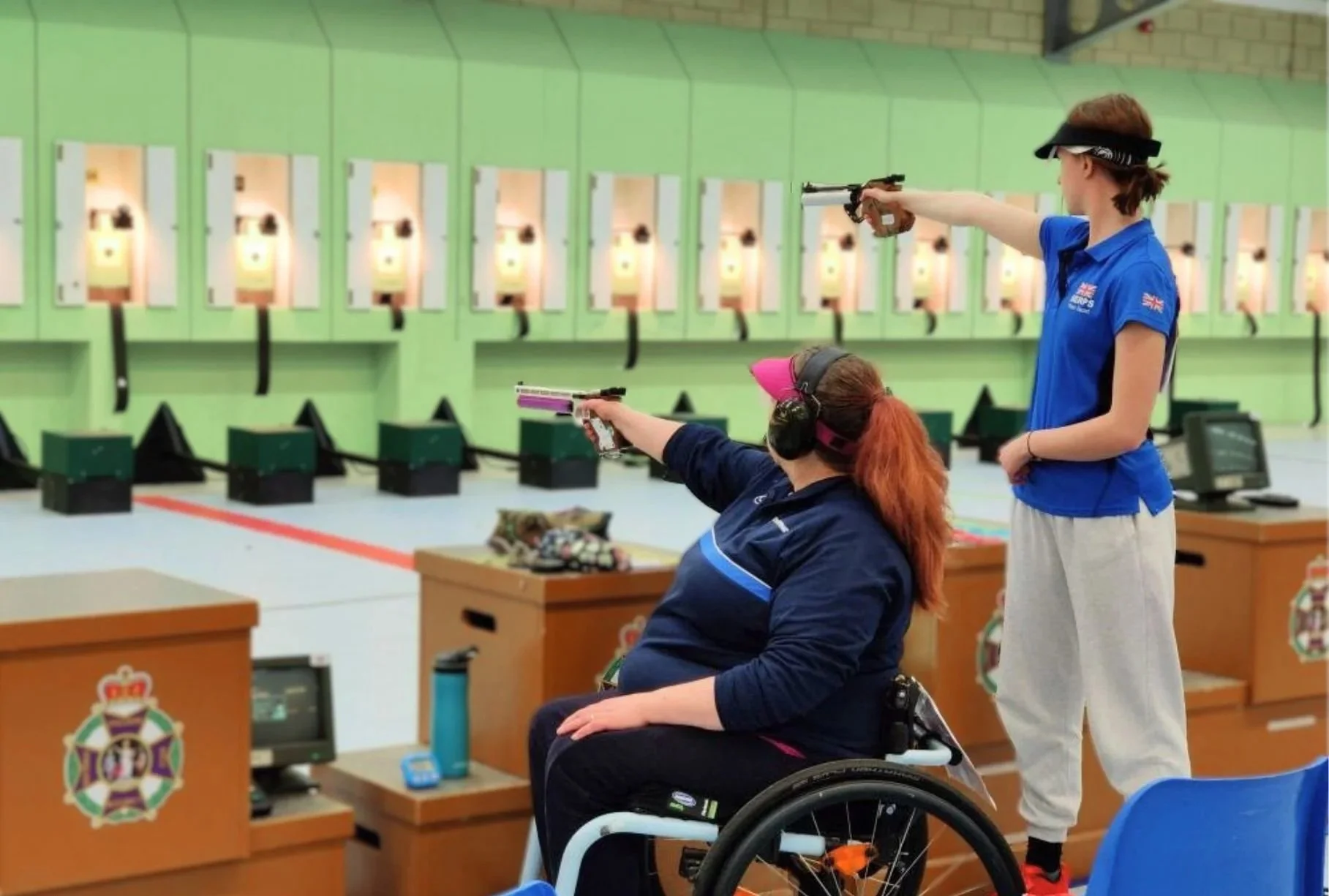 Two people target shooting at NSRA Bisley's events range. One woman is in a wheelchair, aiming a pink pistol, while the other competitor holds a black pistol. The background shows targets on the wall.