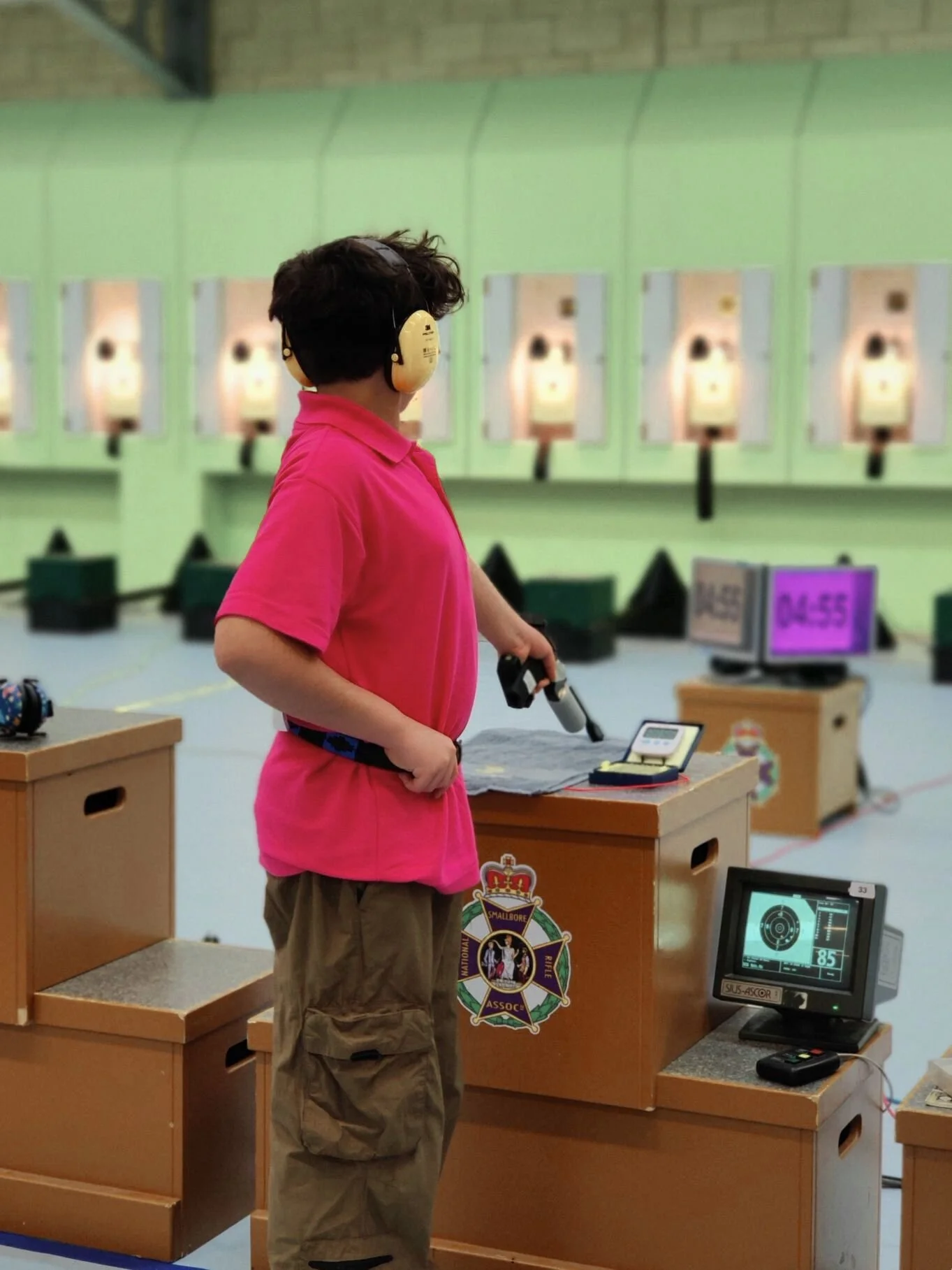 A person wearing a pink shirt, yellow ear protectors and beige cargo pants at a shooting range, on the firing point, holding a pistol, with shooting targets in the background.