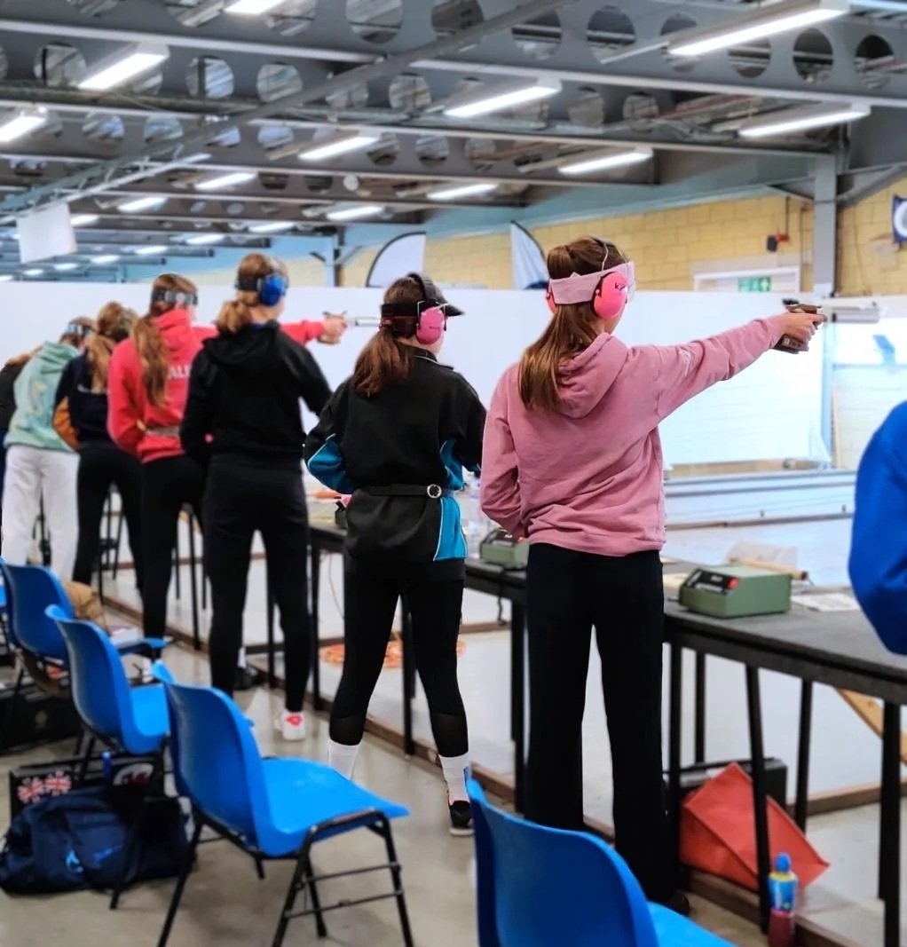 A group of people at NSRA Bisley's Event range, aiming pistols at targets. They are wearing ear protection and casual clothing.
