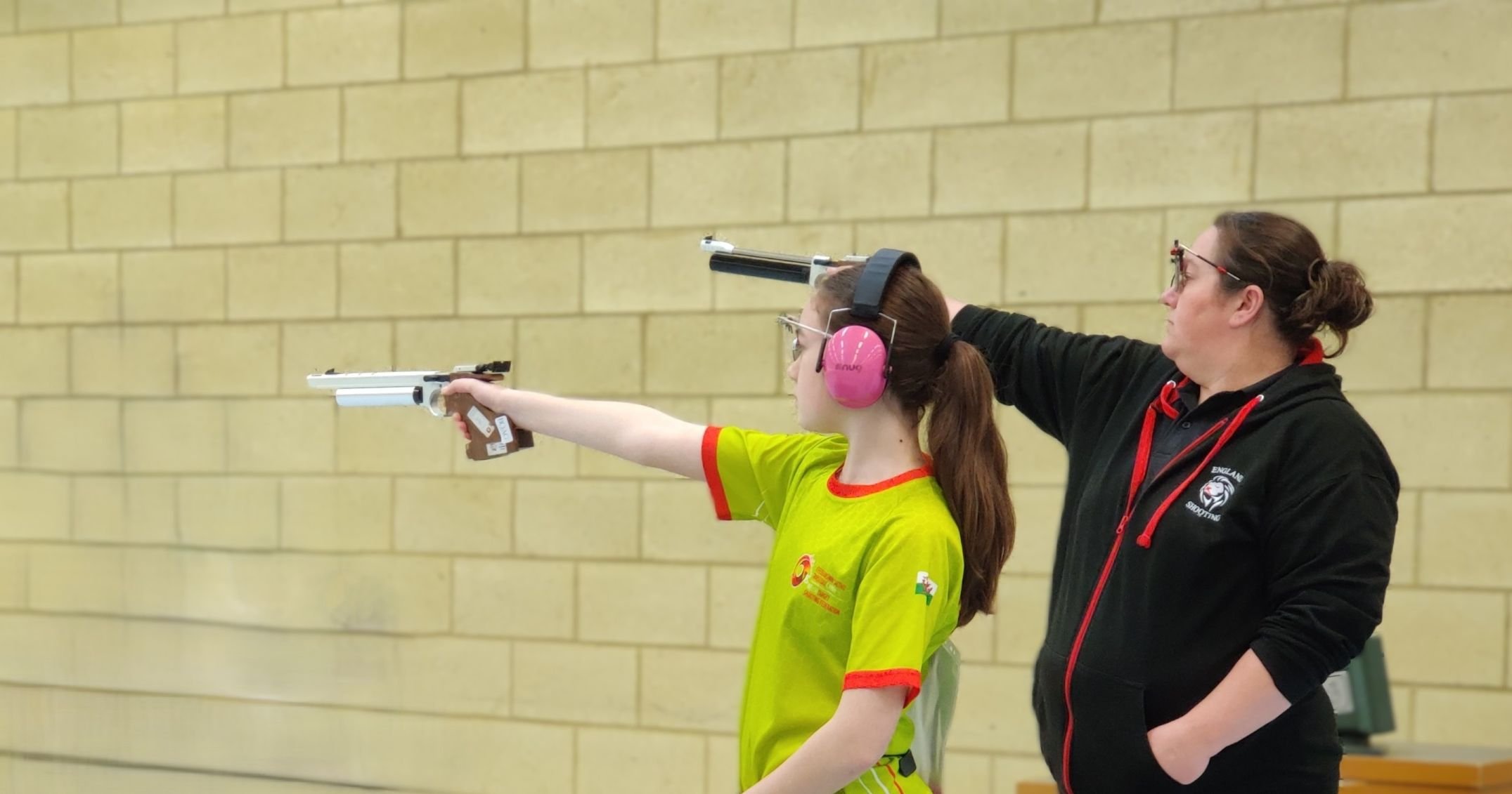 Two people on the firing point taking aim during a pistol competition. One is wearing pink hearing protection, safety glasses, and a yellow sports shirt the other wears a black and red top