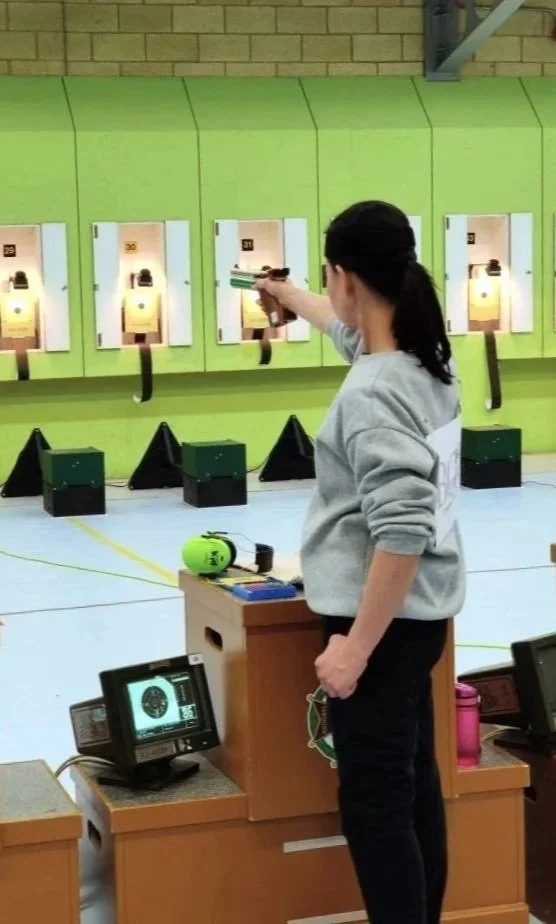 A woman in a gray sweatshirt and black pants at a shooting range, aiming a pistol towards targets on a green wall.