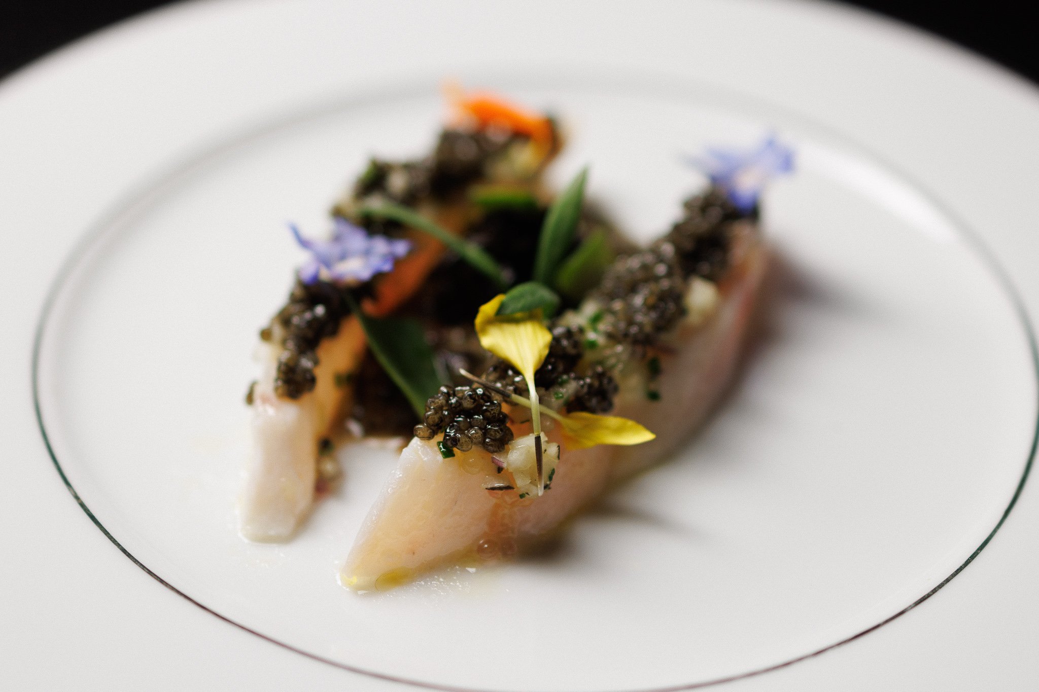 Gourmet plated dish with white fish, caviar, edible flowers, and microgreens on a white plate with a thin black stripe rim.