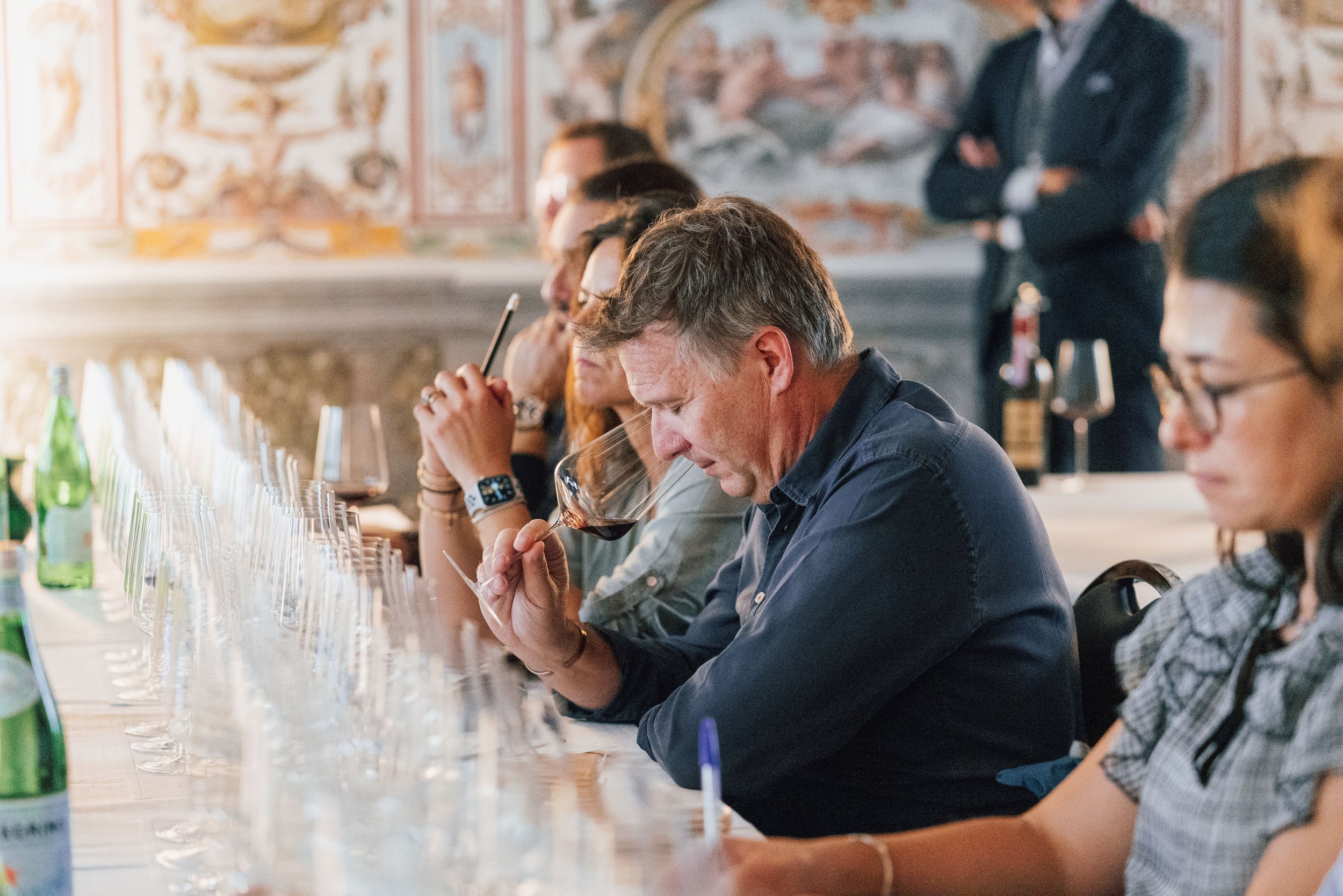 A group of people sit at a long table in a restaurant, tasting glasses of red wine. In the background, a man in a suit stands with his arms crossed. The setting has ornate wall decorations and bottles of water and wine on the table.