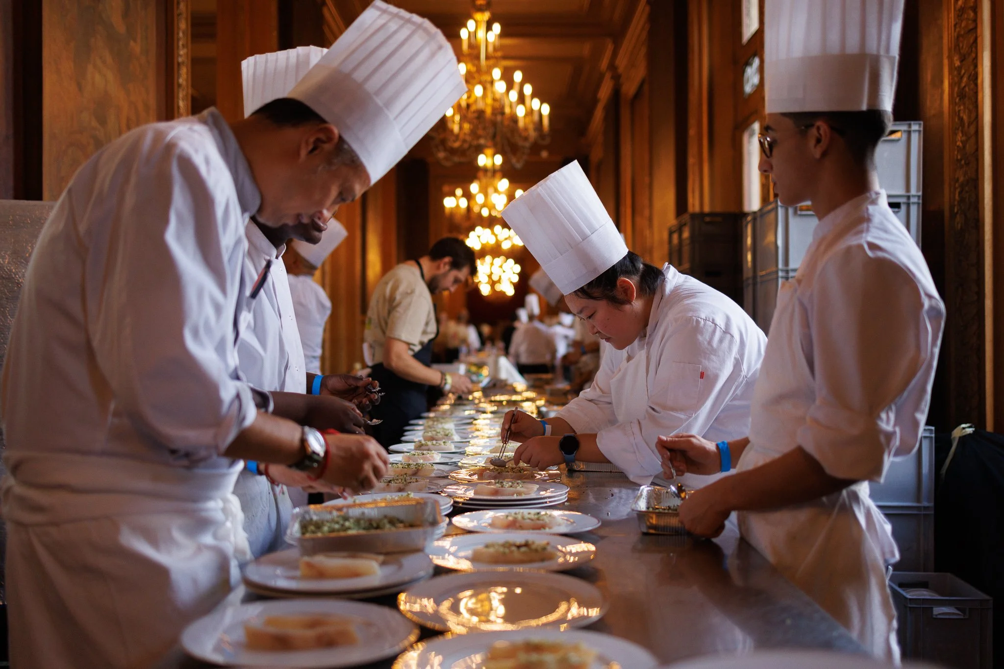 Chefs in white uniforms and tall hats preparing food on a banquet table in an elegant, warmly-lit dining room.