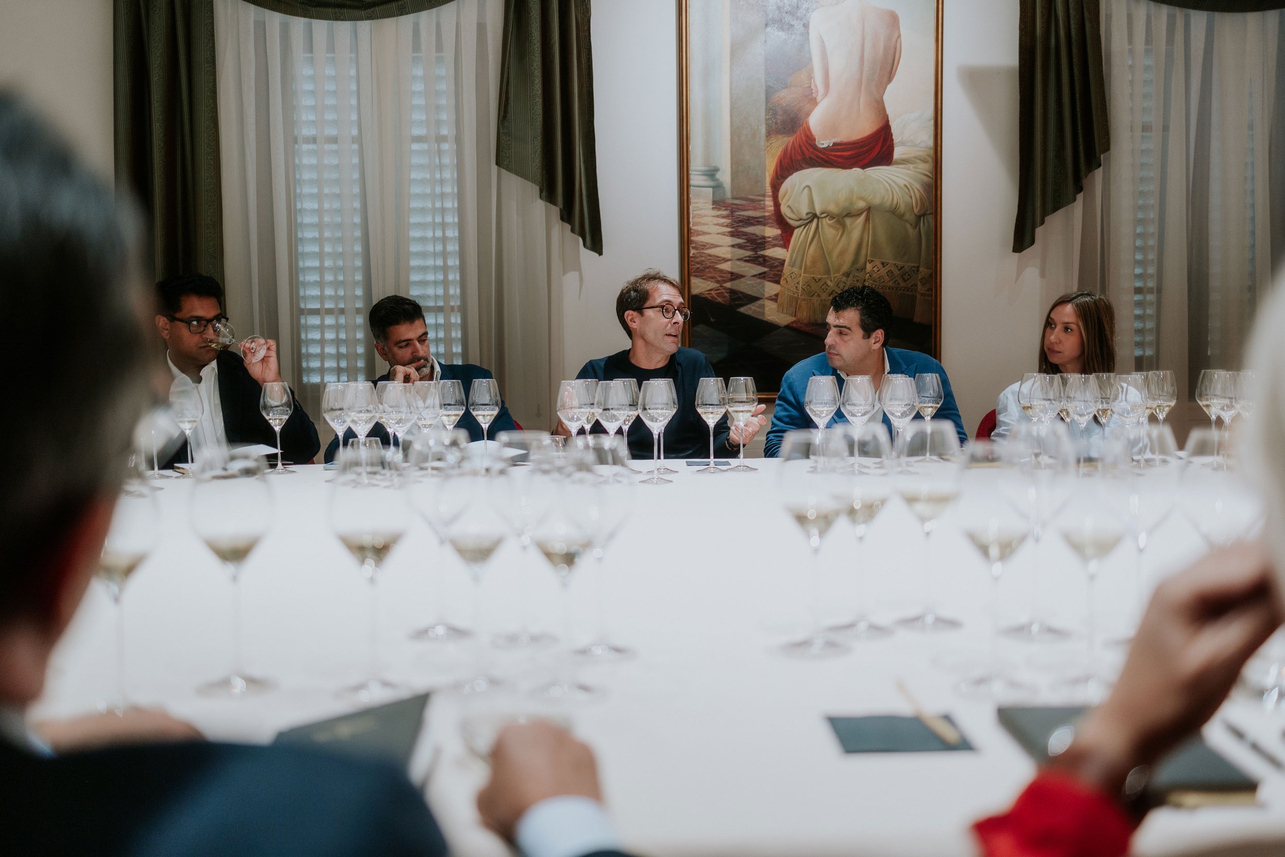 A group of people seated at a long table with multiple empty wine glasses, engaged in conversation, with a large painting of a woman with her back turned on the wall behind them.