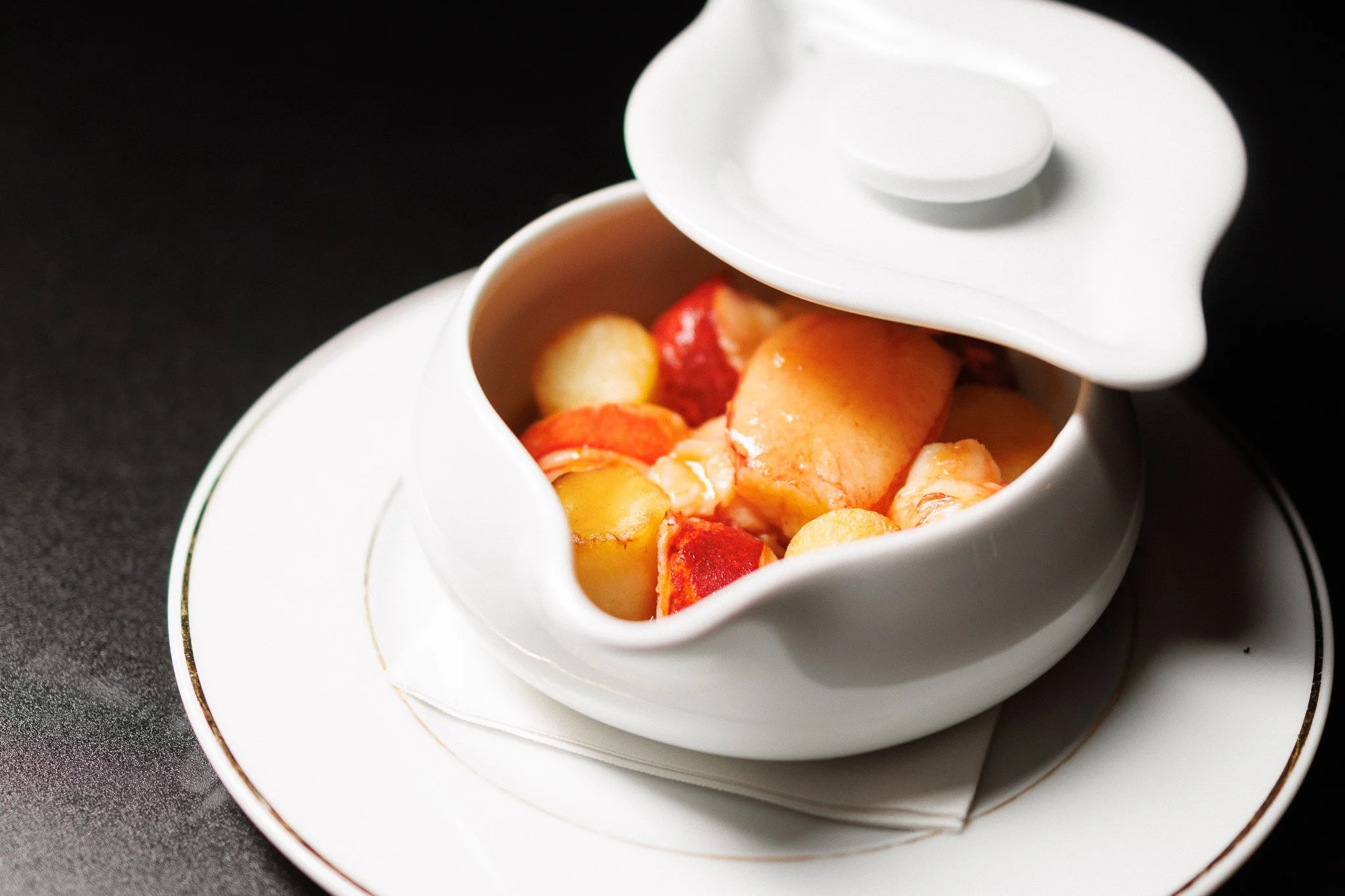 A close-up of a white ceramic teapot with a lid slightly opened, revealing sliced strawberries and peaches inside, placed on a white saucer on a dark surface.