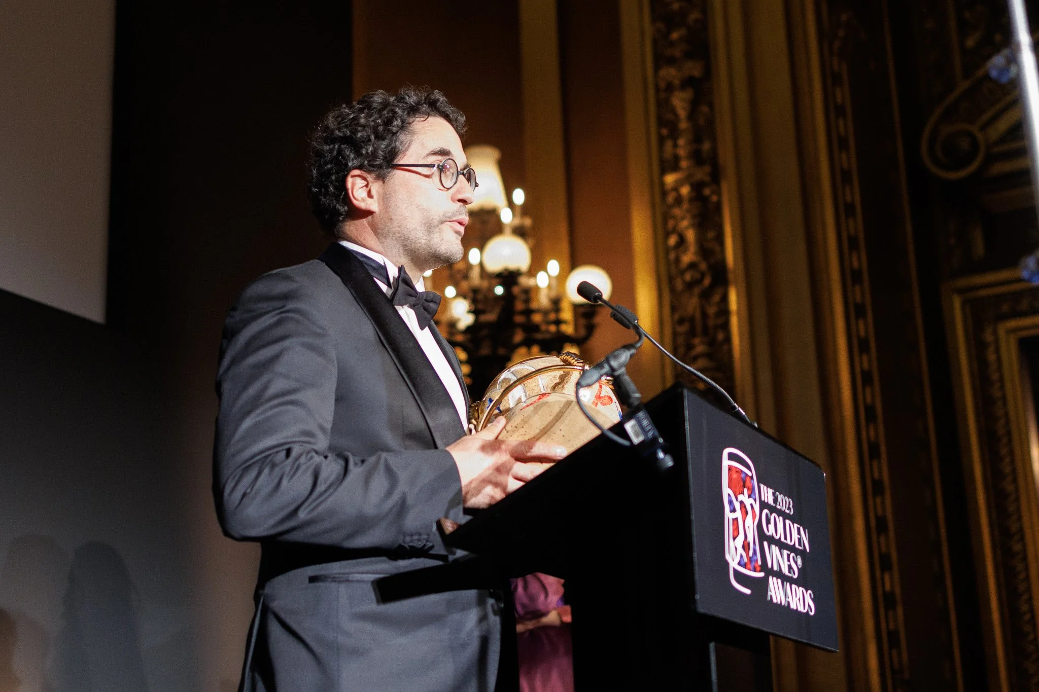 A man in a tuxedo and glasses is speaking at a podium during an awards ceremony inside a decorated hall. He is holding an award and is looking away from the camera.