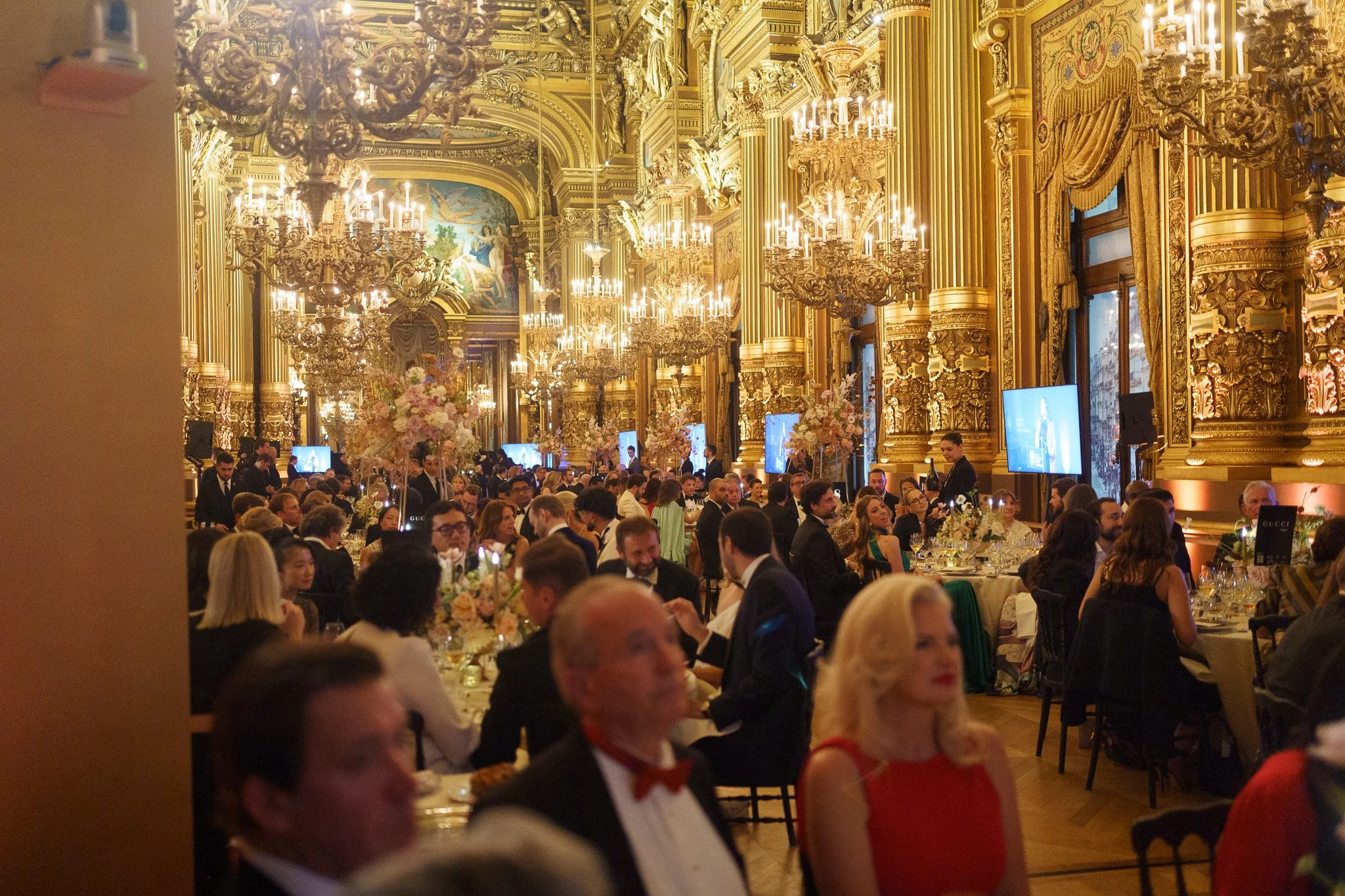A grand banquet hall filled with people dressed in formal attire, with ornate gold decorations, chandeliers, and large floral centerpieces on the tables.