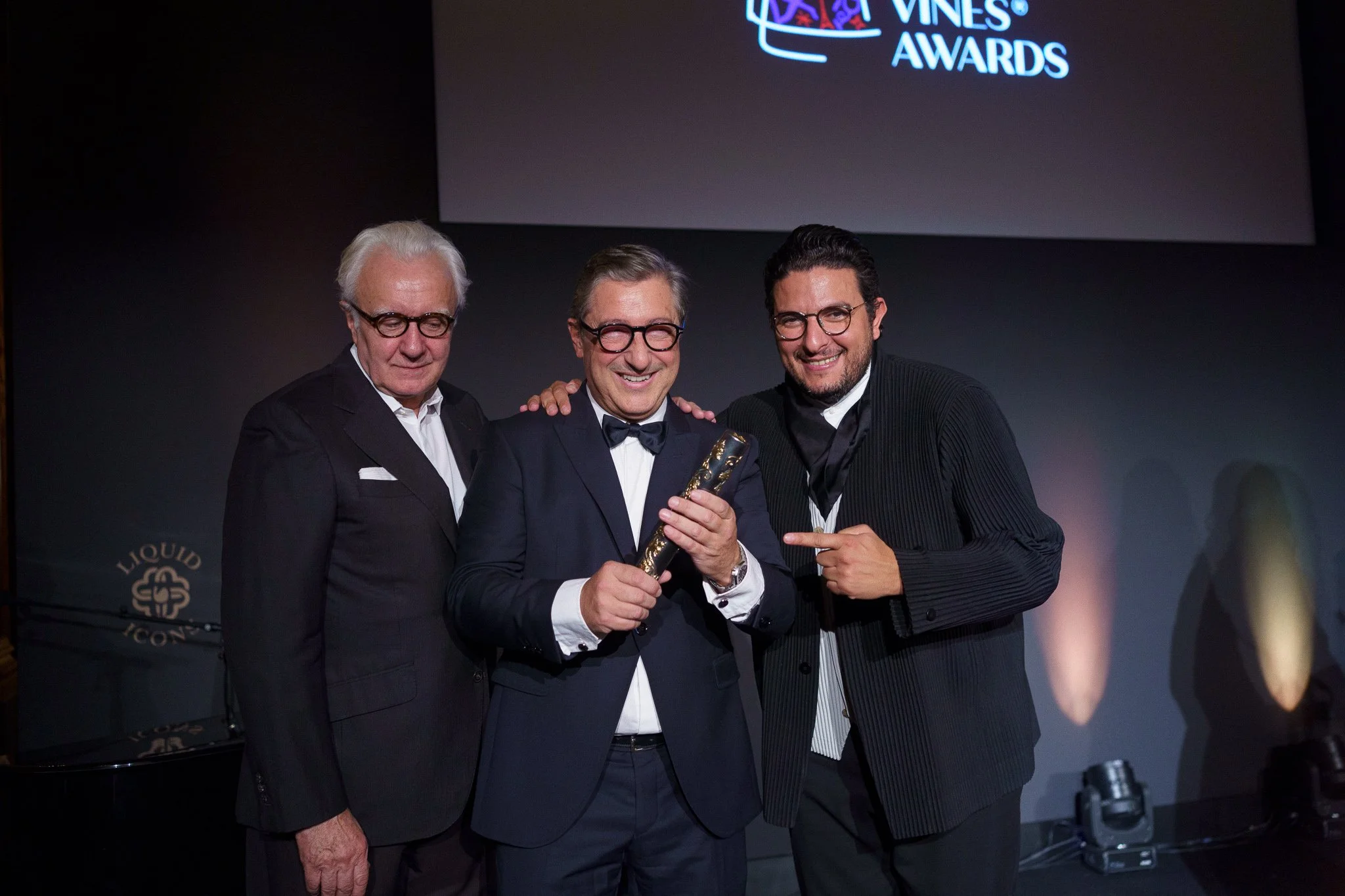 Three men in formal attire celebrating at an awards ceremony, with a large screen behind them displaying the word 'AWARDS' and the Liquor logo.