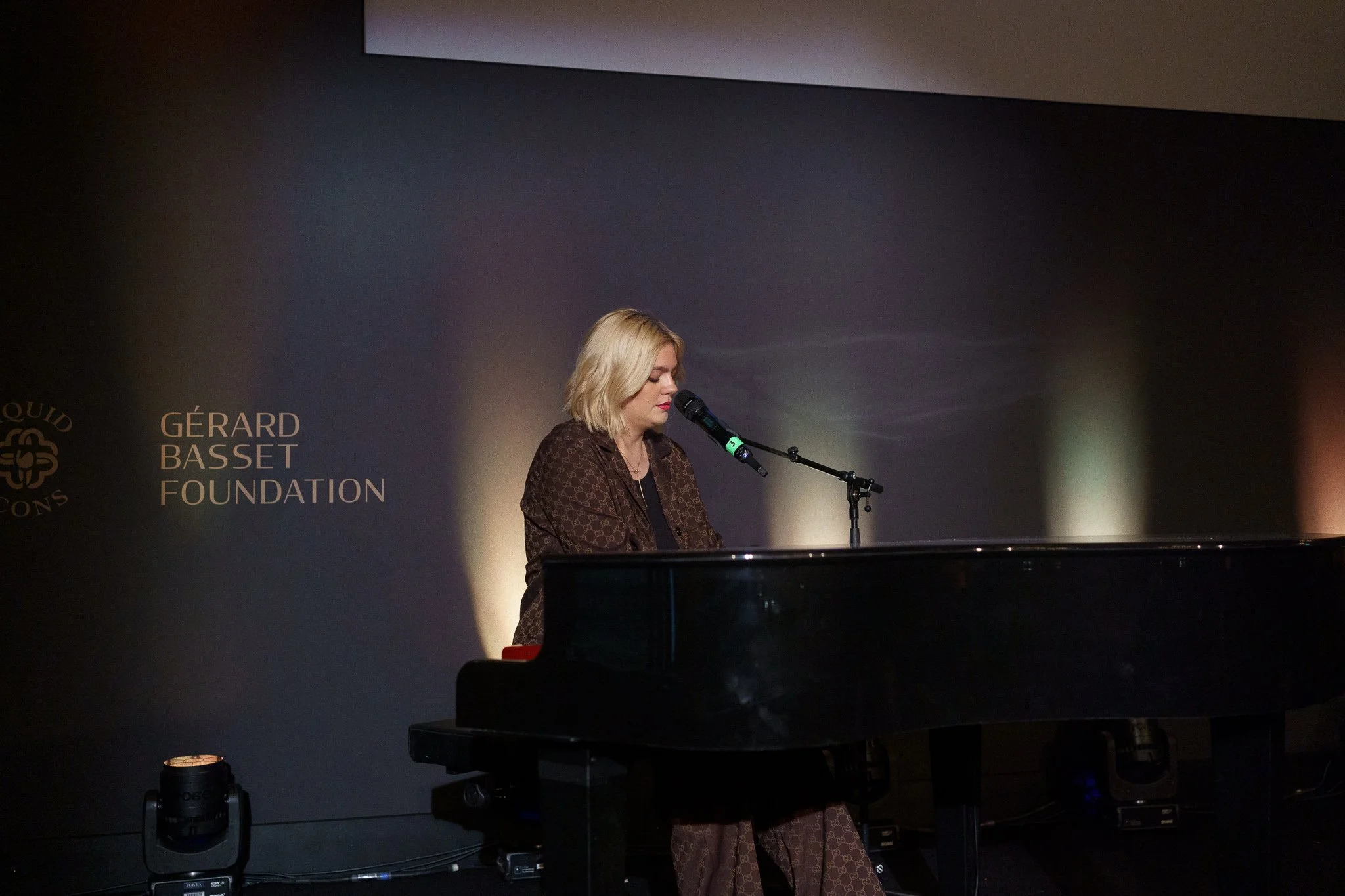 A woman with blonde hair wearing a brown patterned outfit is playing a black grand piano and singing into a microphone in a darkened room. Behind her, a black wall displays the Gérard Basset Foundation logo.