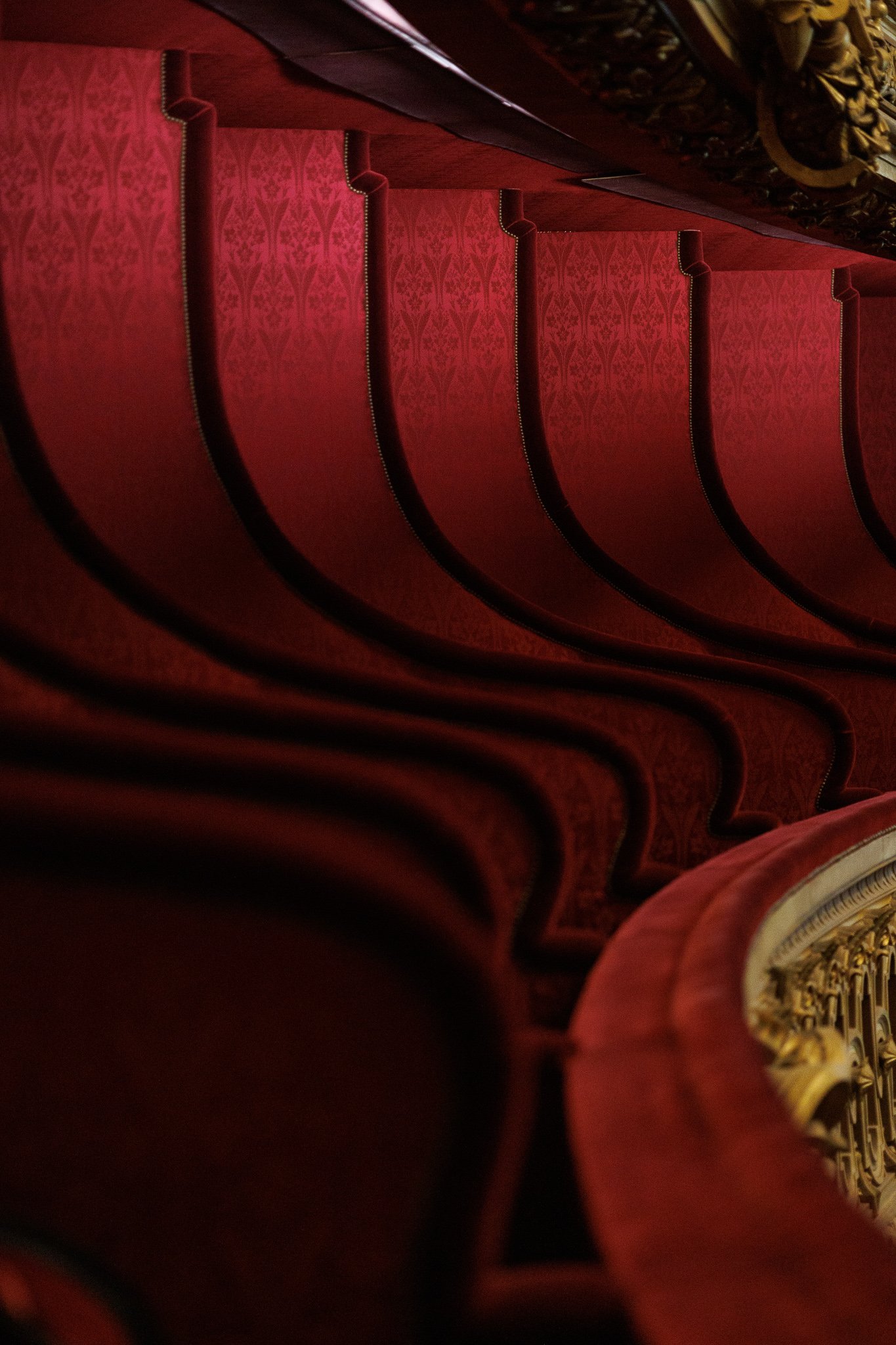 Close-up view of red theater seats with ornate backs, arranged in a row, with a focus on the intricate gold design on the armrest or side of a seat in a theater or opera house.