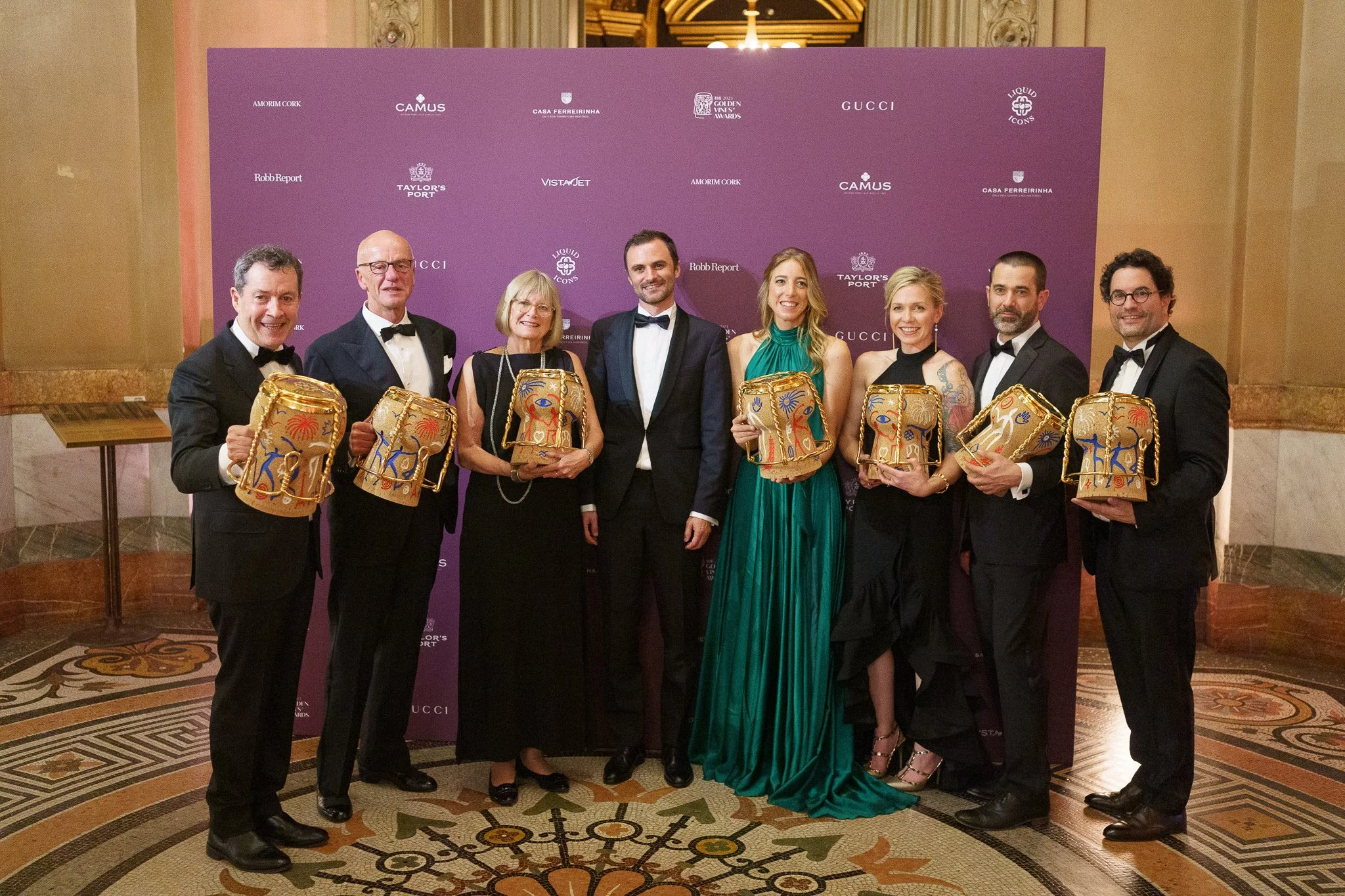 A group of nine people, dressed in formal attire, standing in front of a purple backdrop with logos, holding decorative handbags shaped like heads, at an event in an elegant venue with ornate floors and walls.