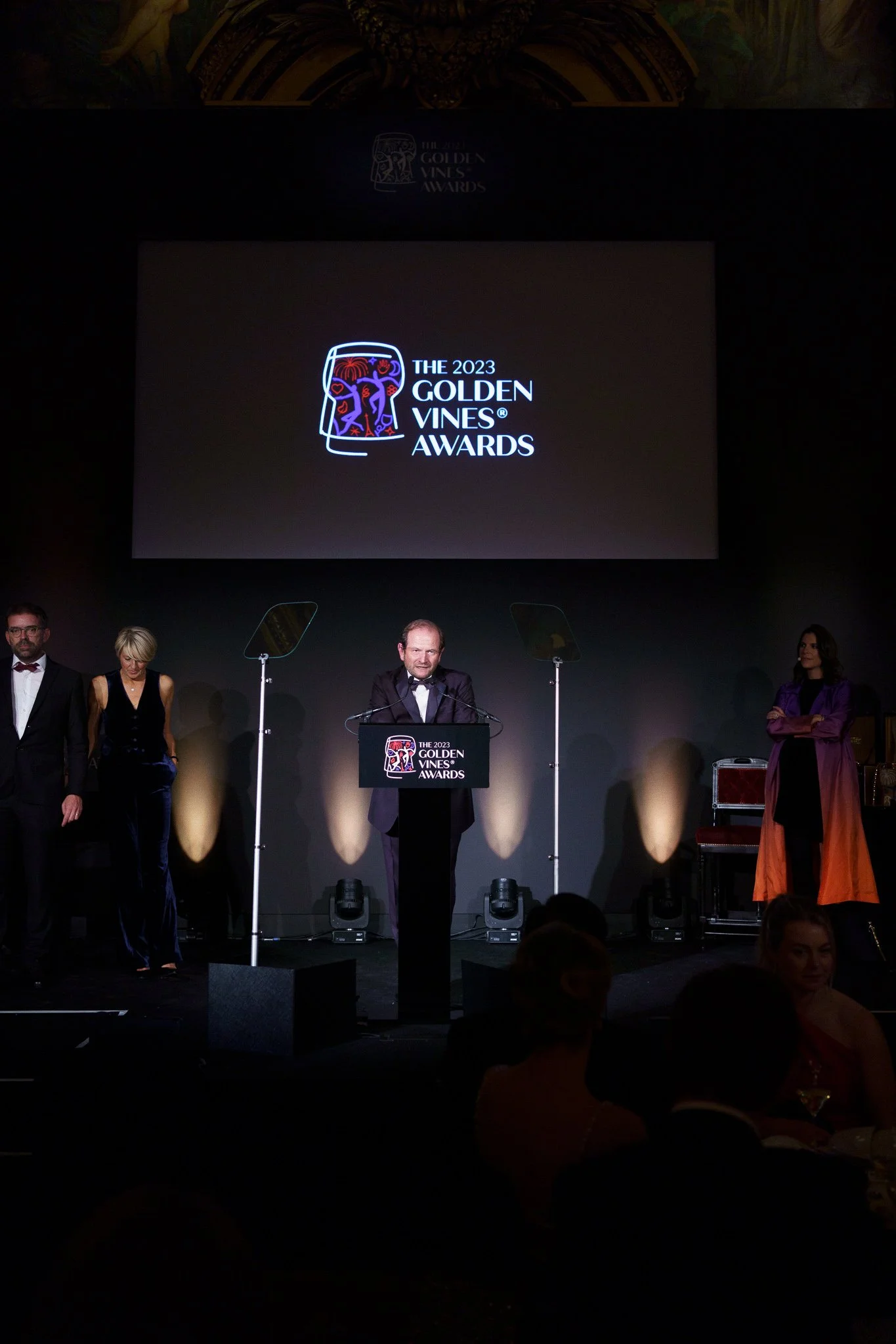 A group of formally dressed people standing on stage at an awards ceremony, with one man speaking at a podium with the logo for the 2023 Golden Vines Awards. There is a large screen behind them displaying the event's logo.