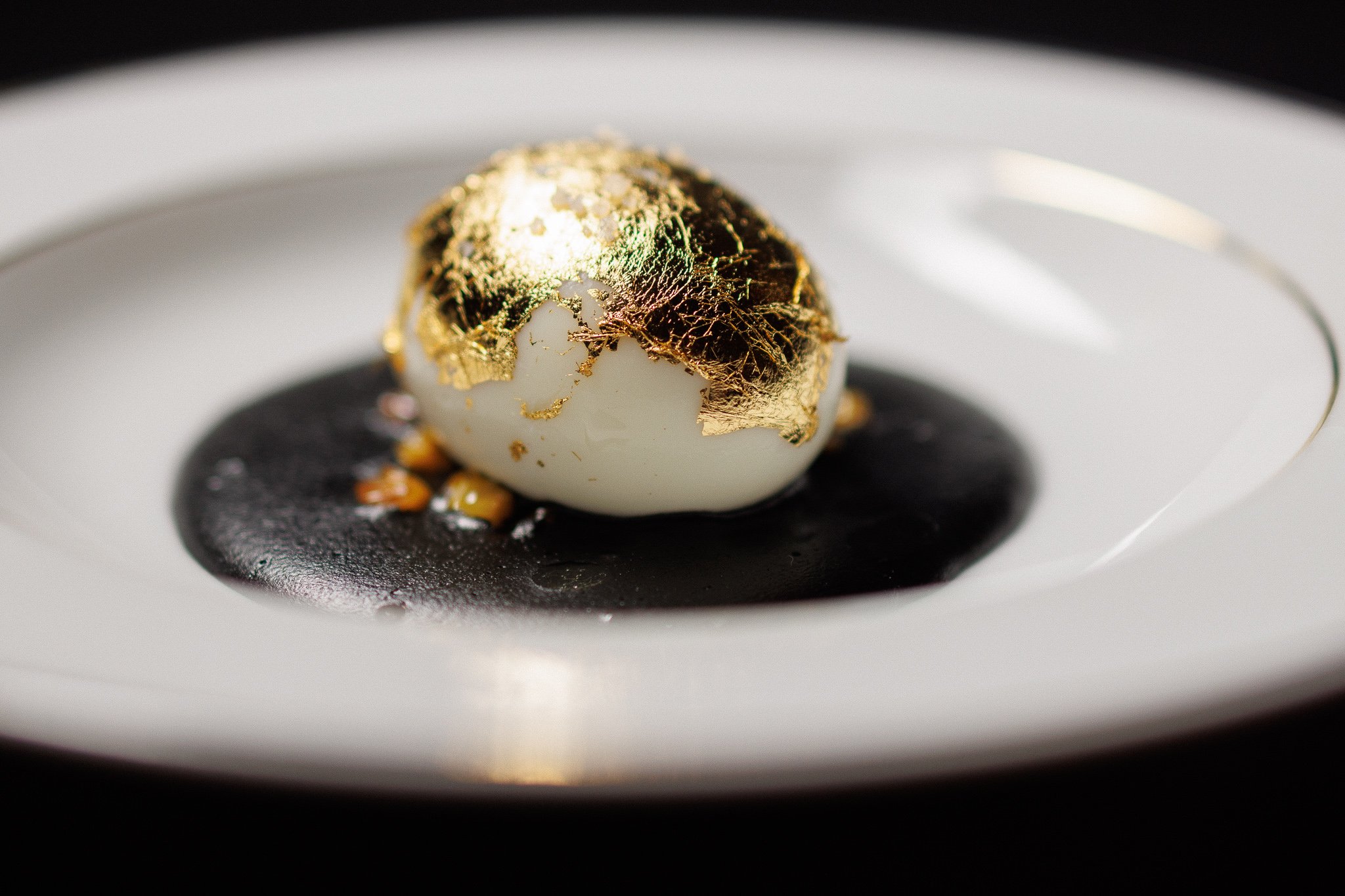 A dessert with a white spherical center topped with gold leaf, served on a white plate with a dark sauce underneath and small garnish around it.
