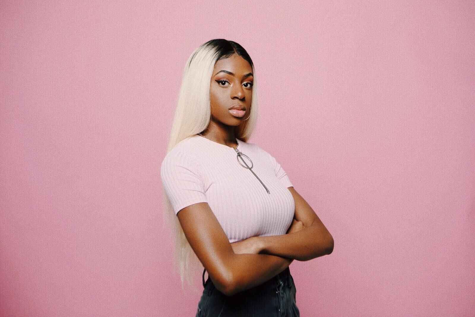 A young woman with long blonde hair, wearing a pink ribbed top with a zipper, stands with crossed arms against a pink background.