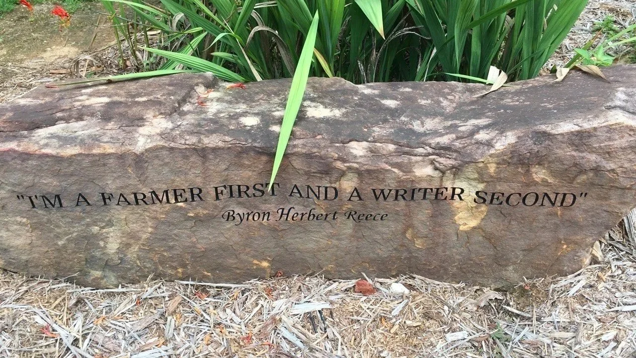 Boulder engraved with "I am a farmer first and a writer second - Byron Herbert Reece" Stone is surrounded by green foilage and natural materials.