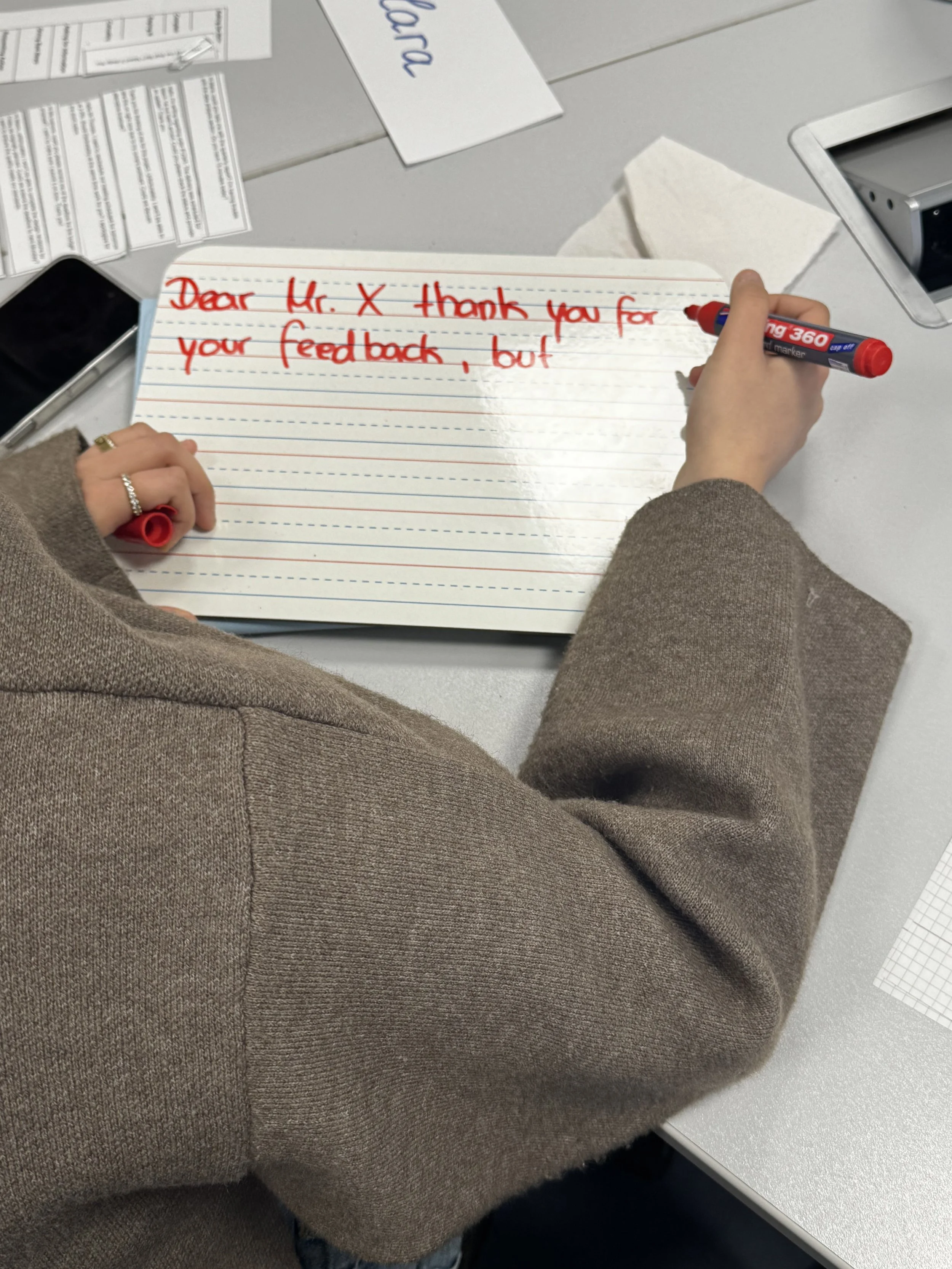 A student wearing a brown blazer is writing on a mini board with a red marker "Dear Mr. X thanks for your feedback, but..."