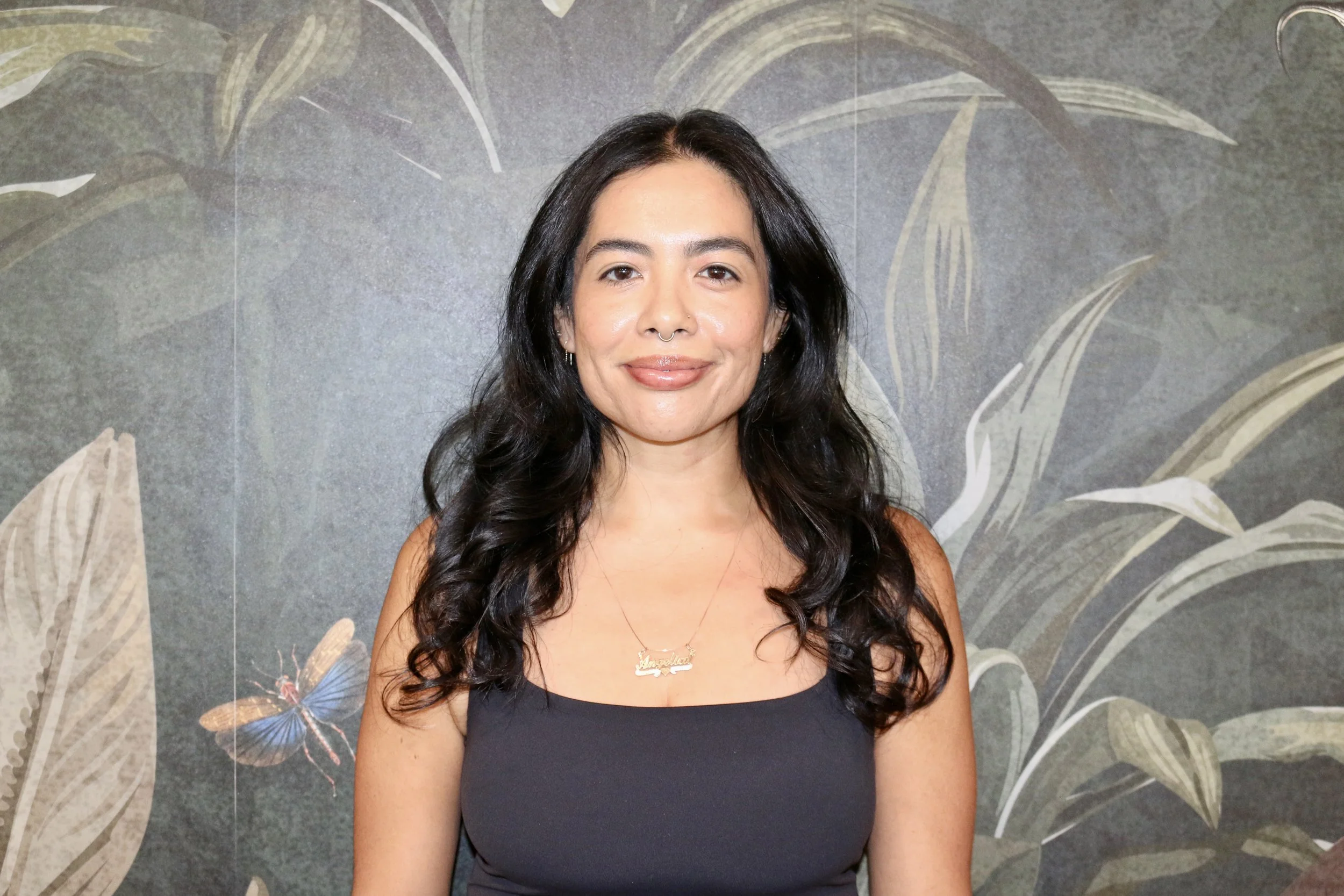 A woman with long, curly dark hair, wearing a black tank top and a necklace, standing in front of a decorative wall with a leaf and butterfly design.