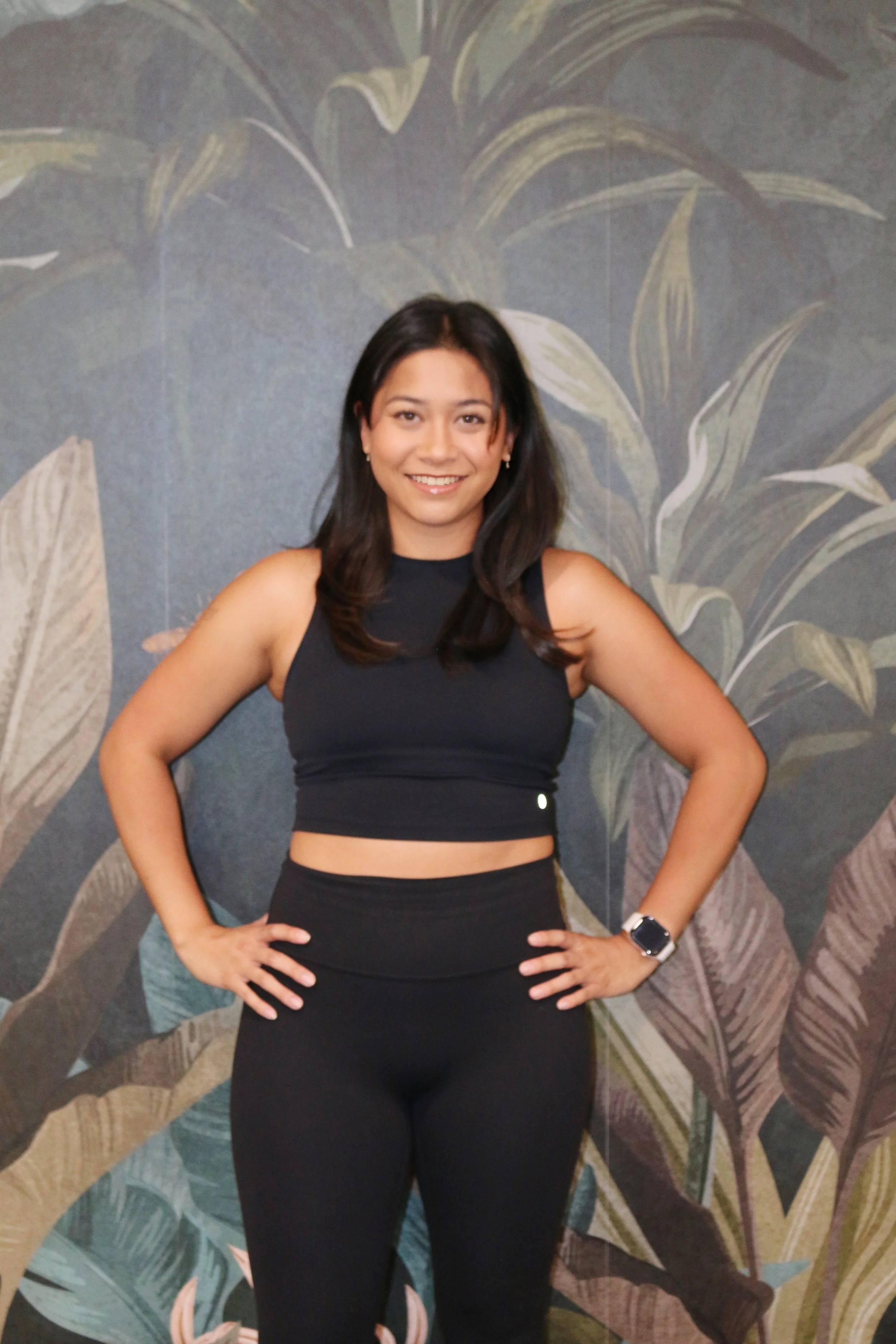 A woman with dark hair, wearing a black sleeveless crop top and black leggings, smiling and standing in front of a leafy, tropical-patterned wall.