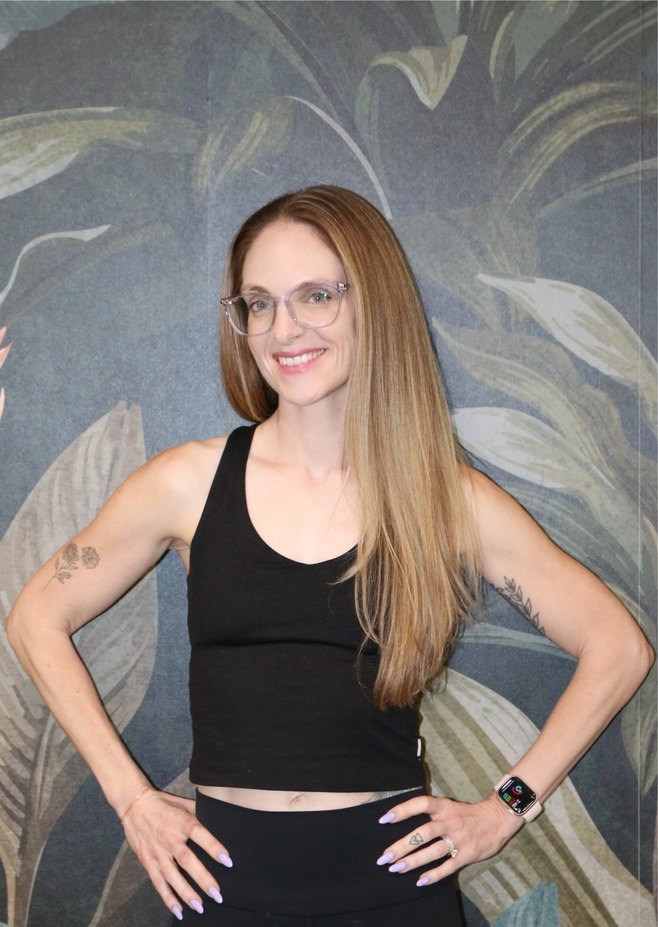 A young woman with long, straight, light brown hair, wearing glasses, a black sleeveless top, and black pants, stands with her hands on her hips smiling in front of a dark wall with large, abstract leaf patterns.