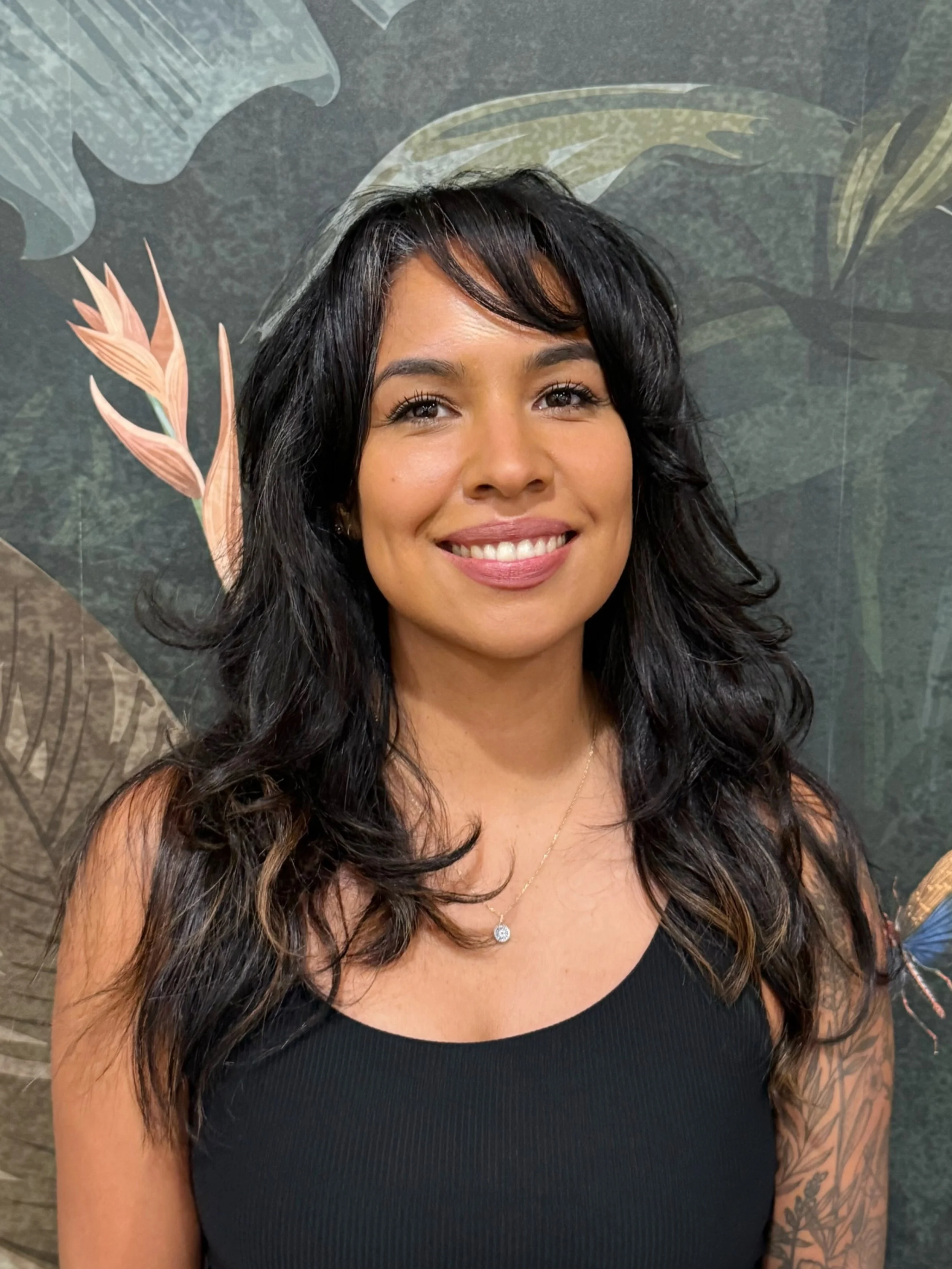 A woman with long, wavy black hair smiling, wearing a black sleeveless top and a delicate necklace, standing in front of a dark botanical mural background.