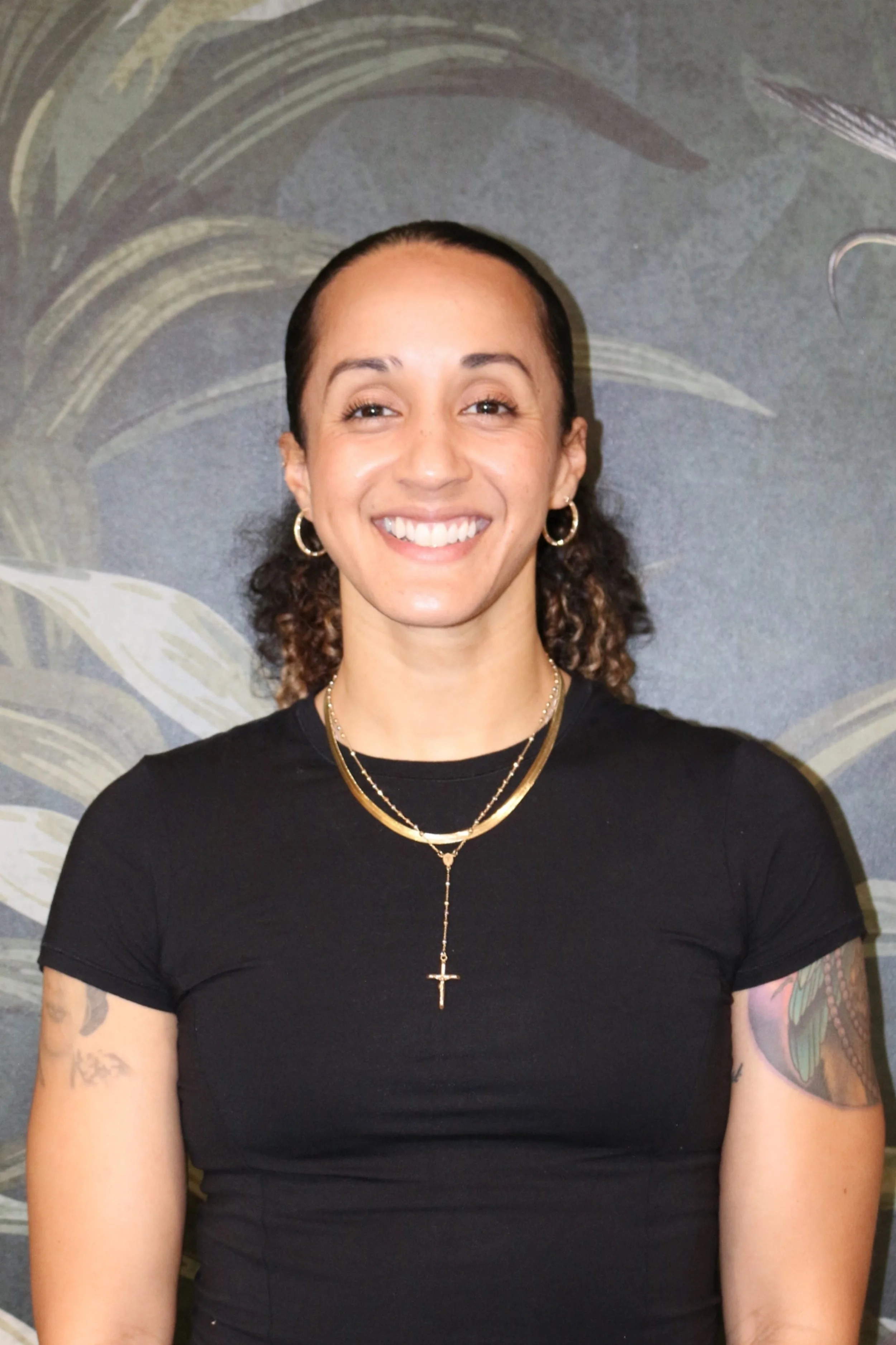 A smiling woman with curly hair wearing a black t-shirt, gold jewelry, and tattoos on her arms, standing in front of a decorative background with leaf patterns.