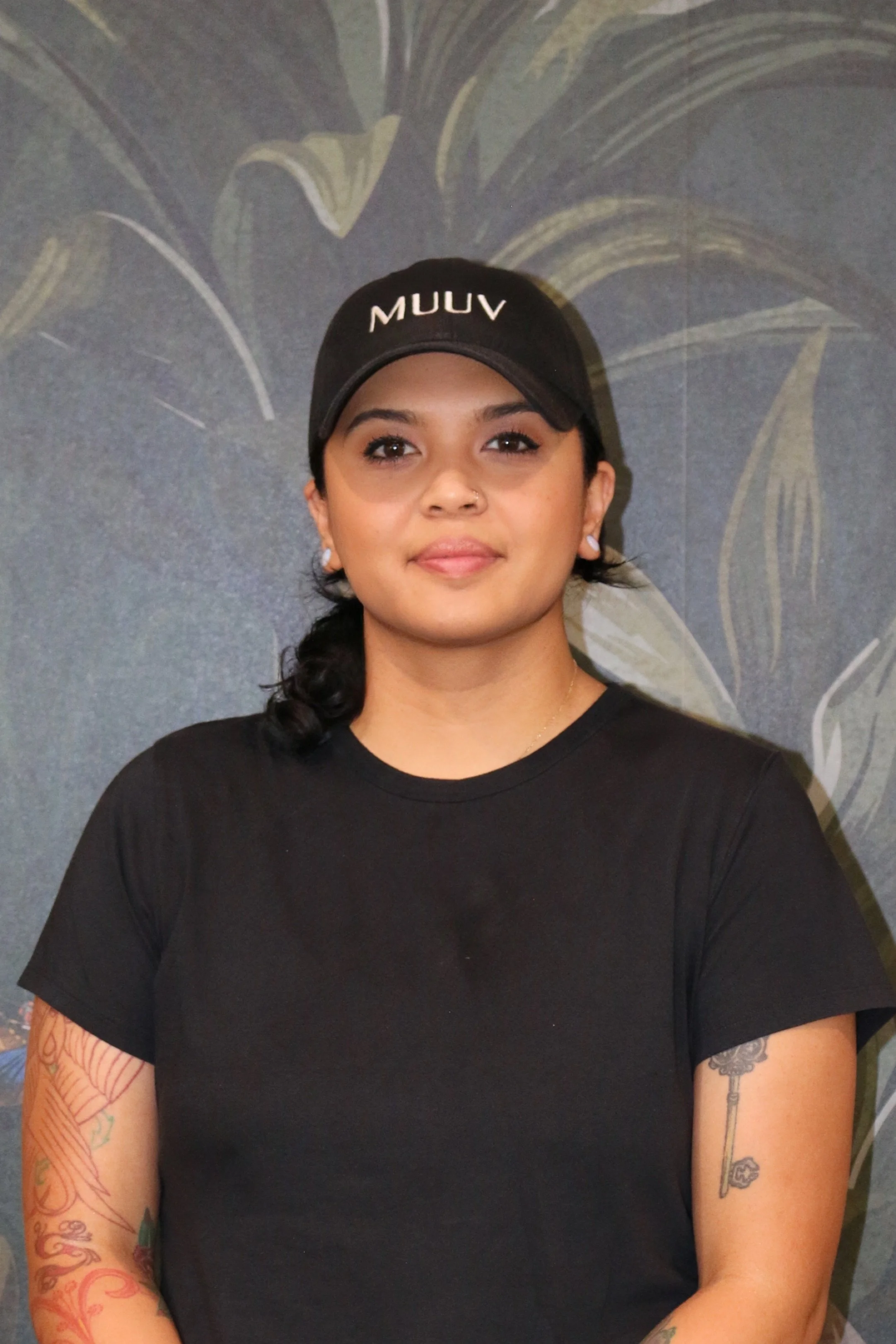 A young woman with medium skin tone, black hair, and tattoos on both arms, wearing a black t-shirt and a black cap with 'MUUV' written on it, standing in front of a patterned wall background.