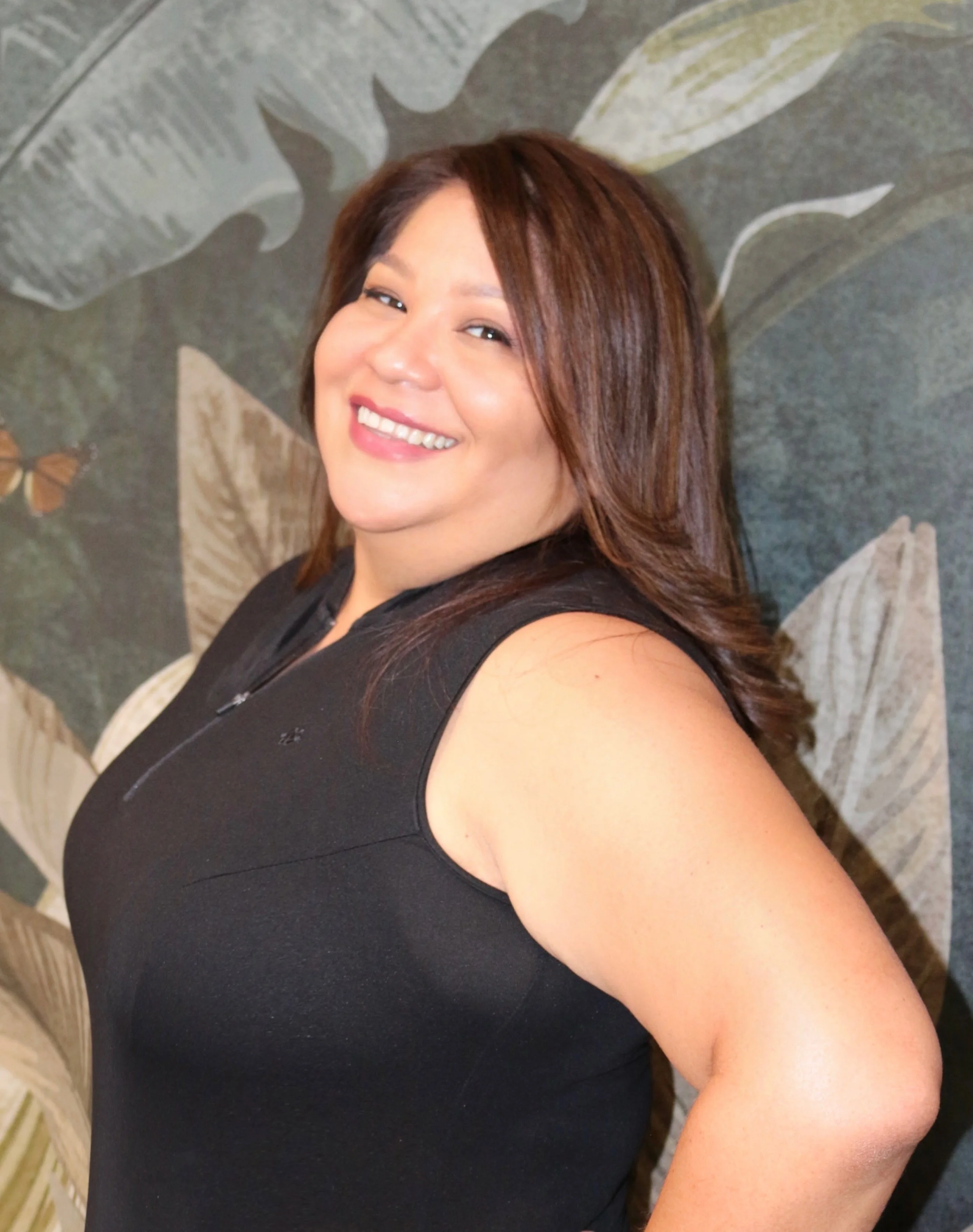 A woman with brown hair smiling at the camera, wearing a sleeveless black top, standing in front of a leafy patterned background.