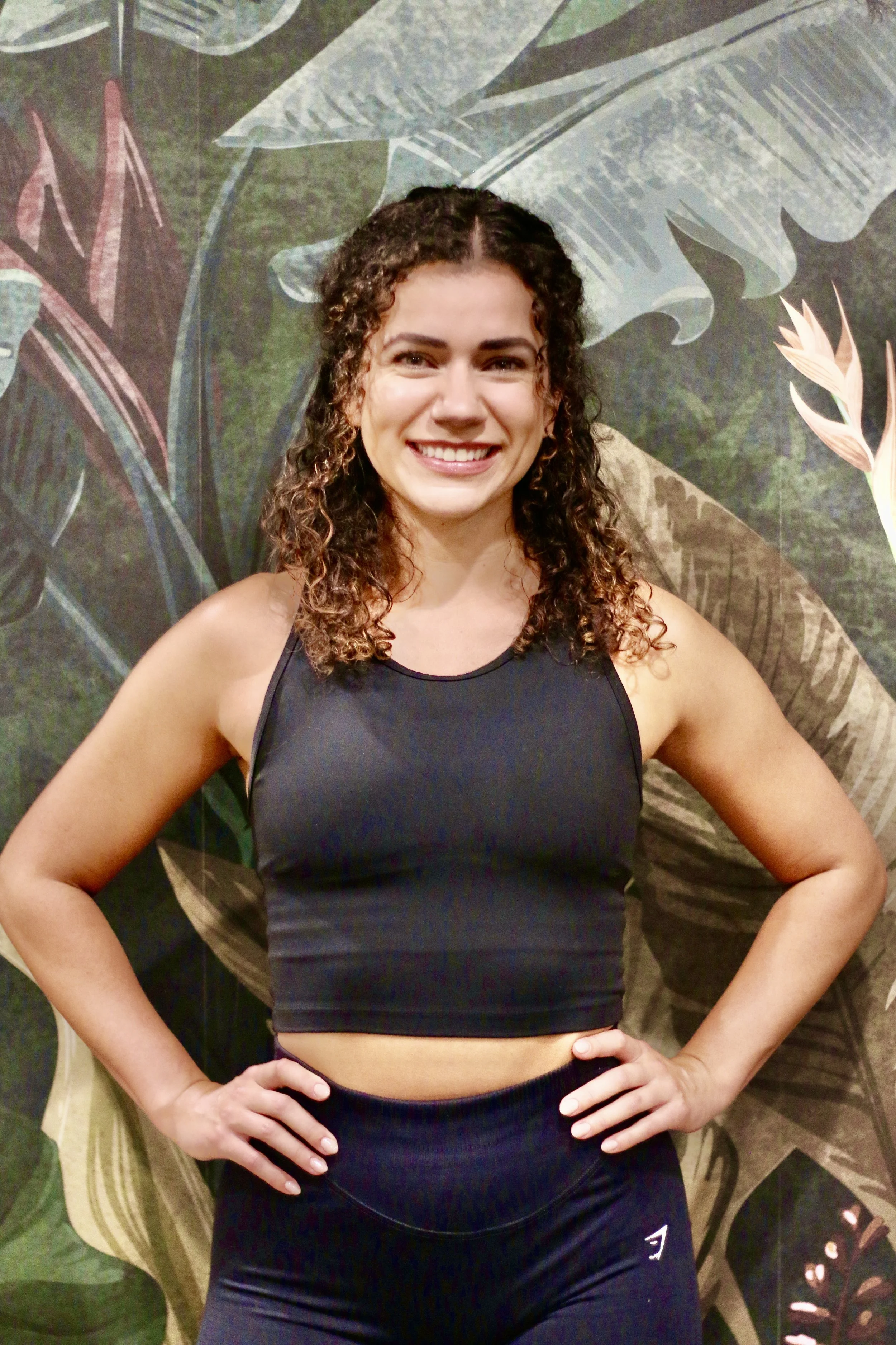 A young woman with curly brown hair wearing a black athletic tank top and black leggings, smiling and standing with her hands on her hips against a tropical-themed wallpaper background.
