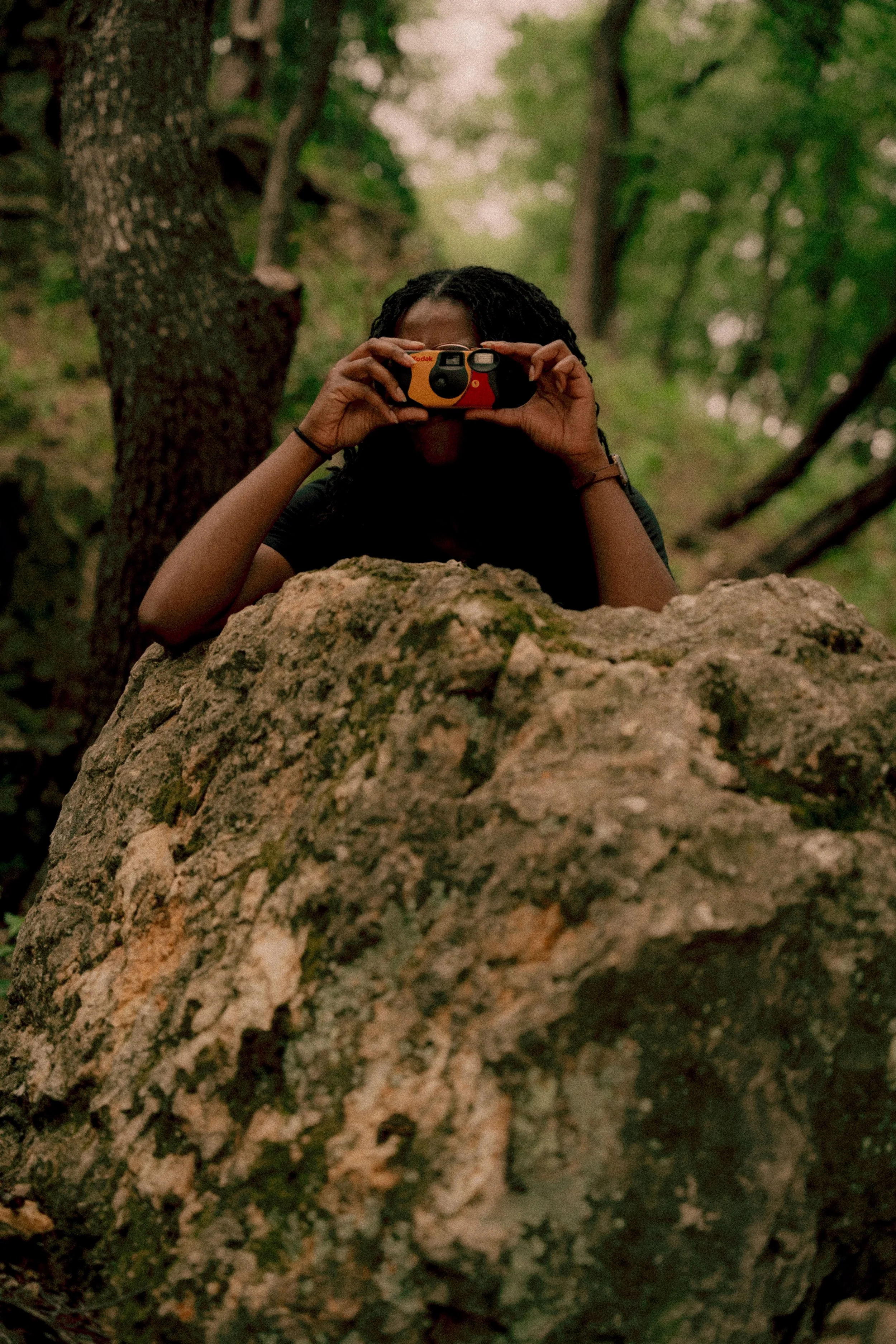 Person lying behind a large rock in a green forest, taking a photo with a camera.