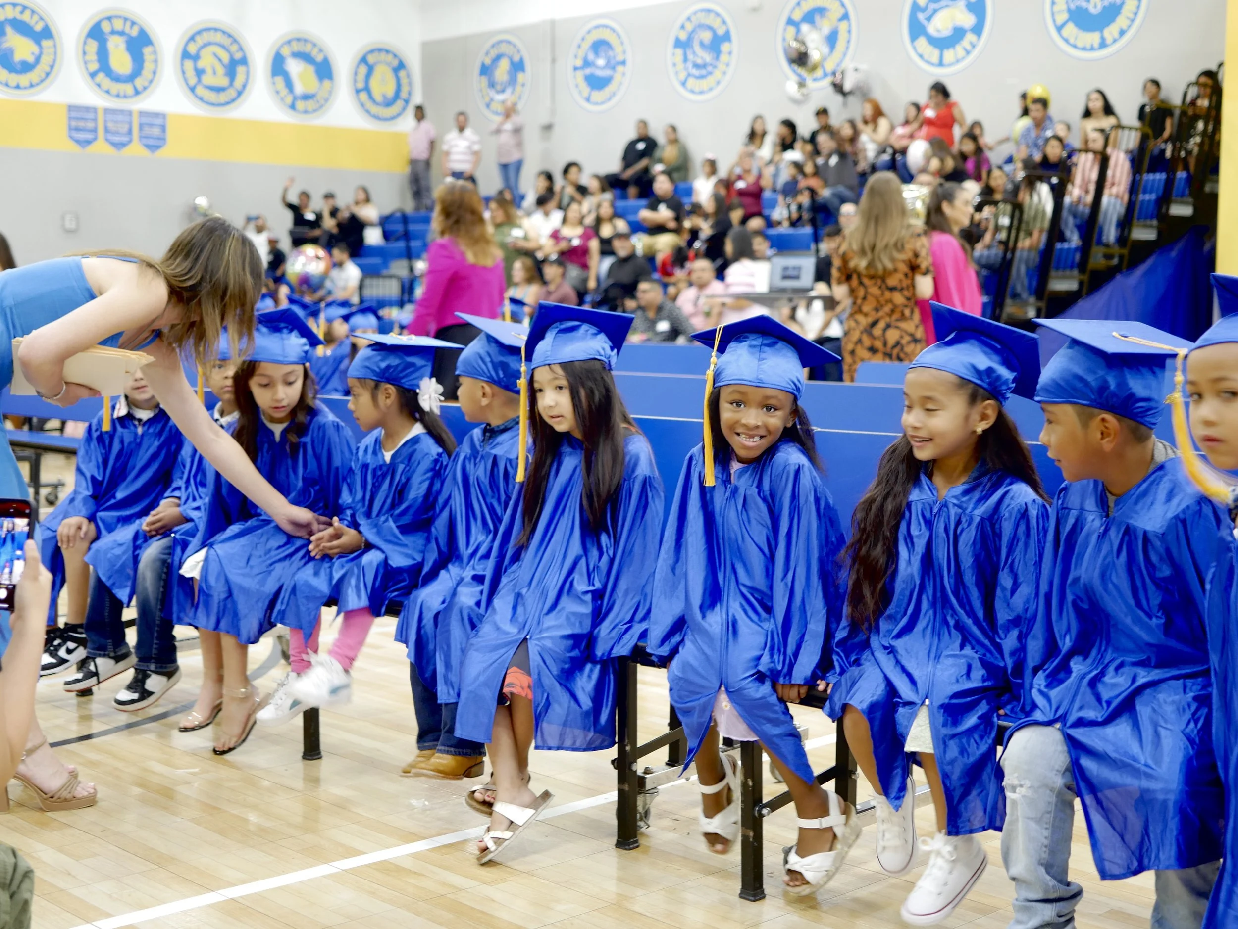 Young children in blue caps and gowns sitting in a row at a graduation ceremony in a gymnasium, with family members and spectators in the background.