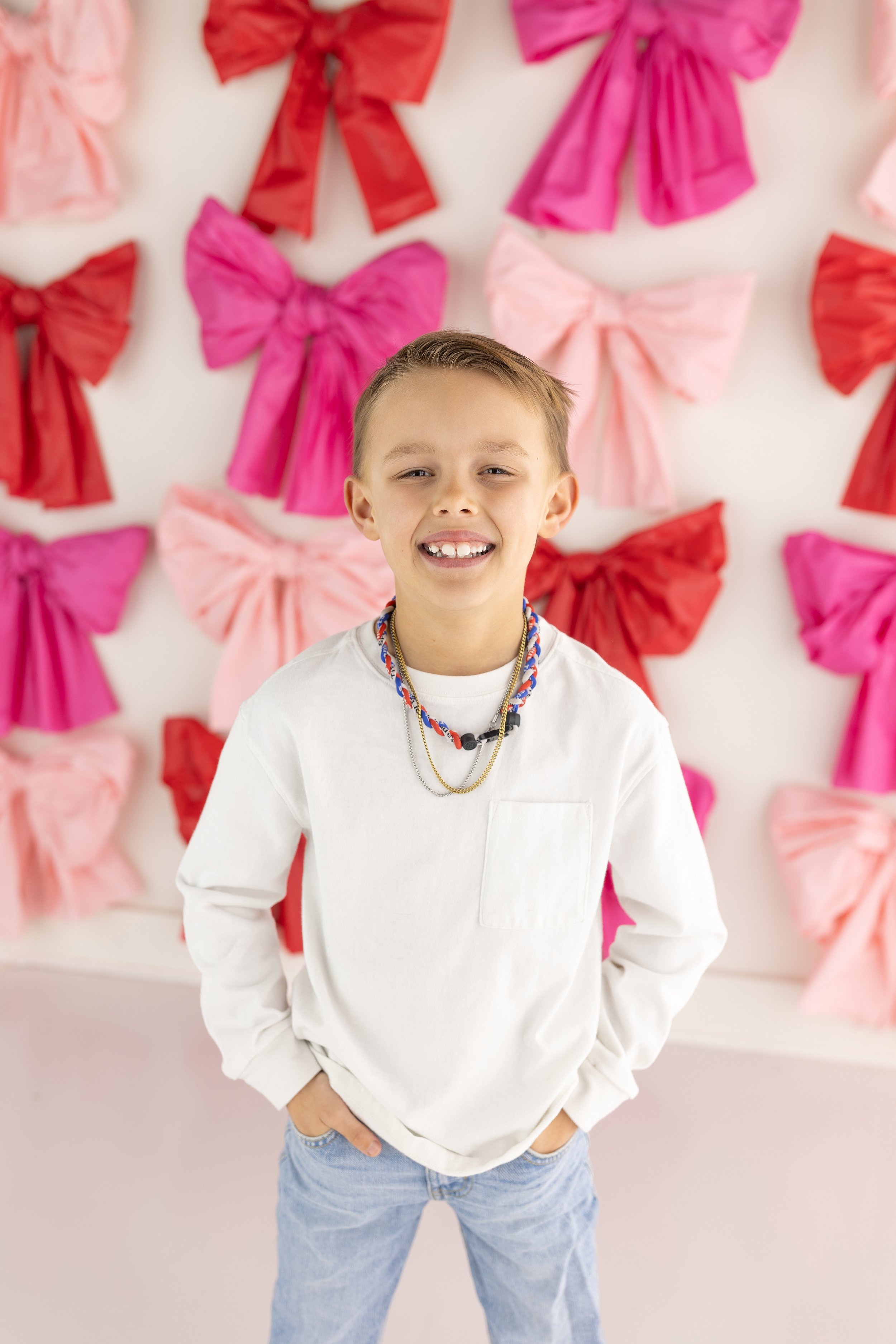 boy standing in front off bow wall for valentines set. boy is wearing white shirt and denim jeans. best studio in northwest arkansas