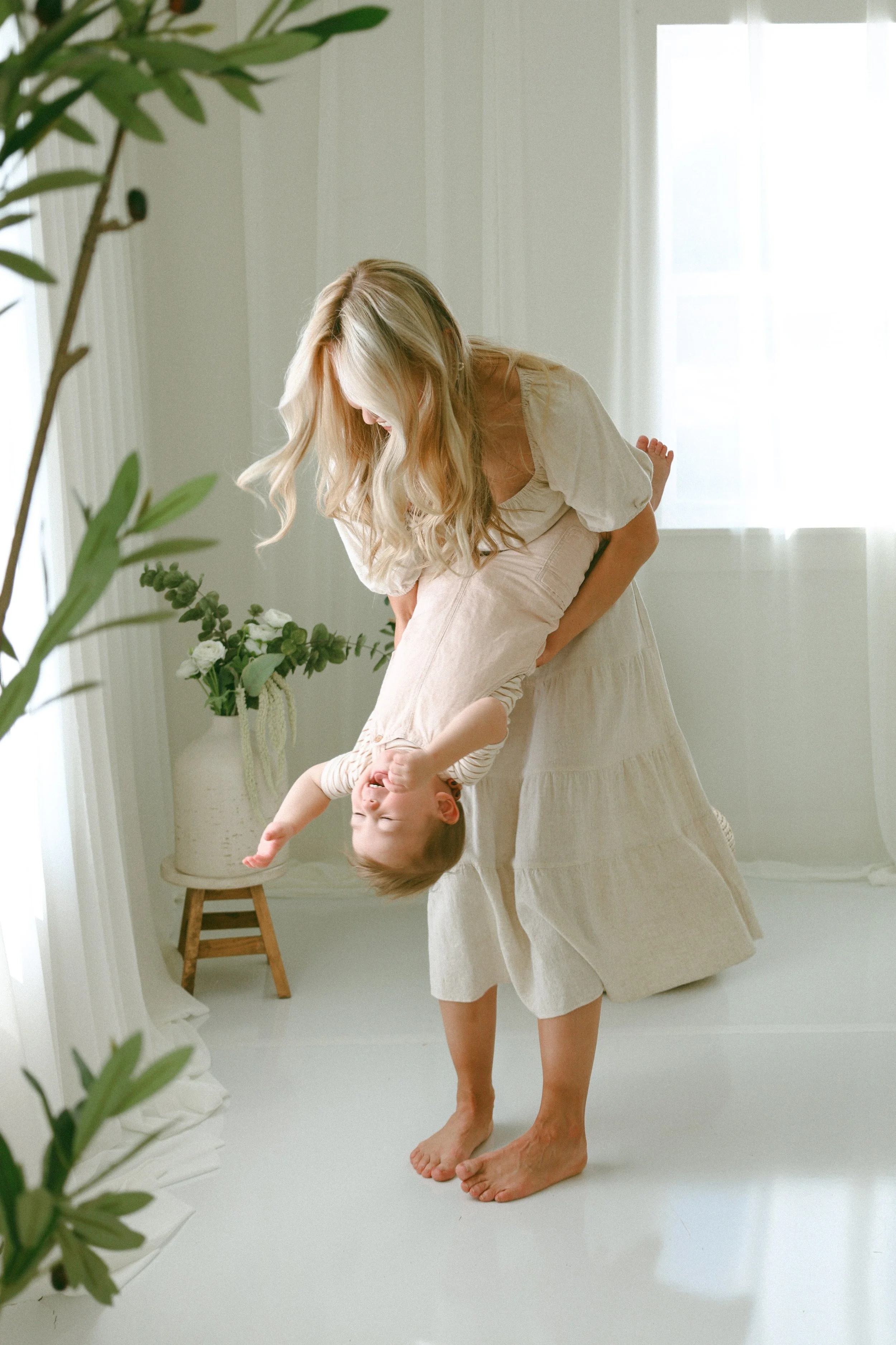 Mother joyfully swings her smiling toddler upside down in a bright, airy white studio with soft natural light and neutral décor at The Studio on White Street in Northwest Arkansas. This candid lifestyle motherhood portrait captures the authentic conn