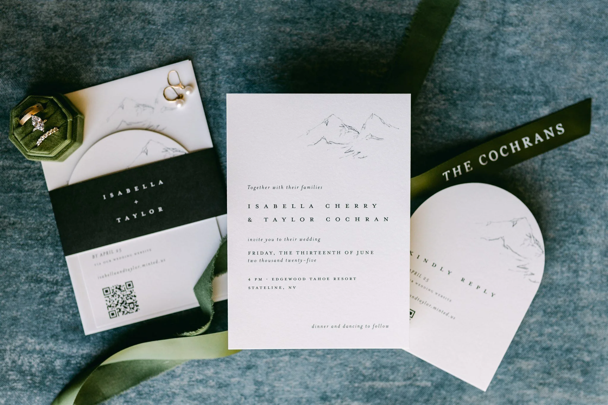 Lake Tahoe wedding invitation suite with mountain sketch illustration, sage green ribbon, green velvet ring box, gold wedding rings, and pearl earrings on a blue linen background — Edgewood Tahoe Resort wedding