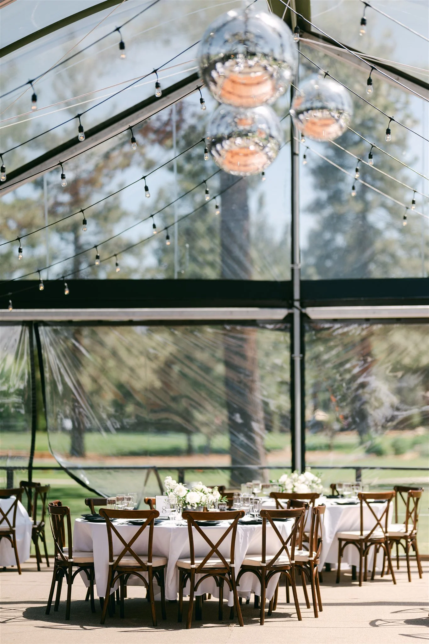Clear tent wedding reception setup at Edgewood Tahoe Resort with cluster disco balls, Edison string lights, white linen round tables, and cross-back wood chairs overlooking Lake Tahoe pines — Sierra Lighting and Mountain Vibes Rentals
