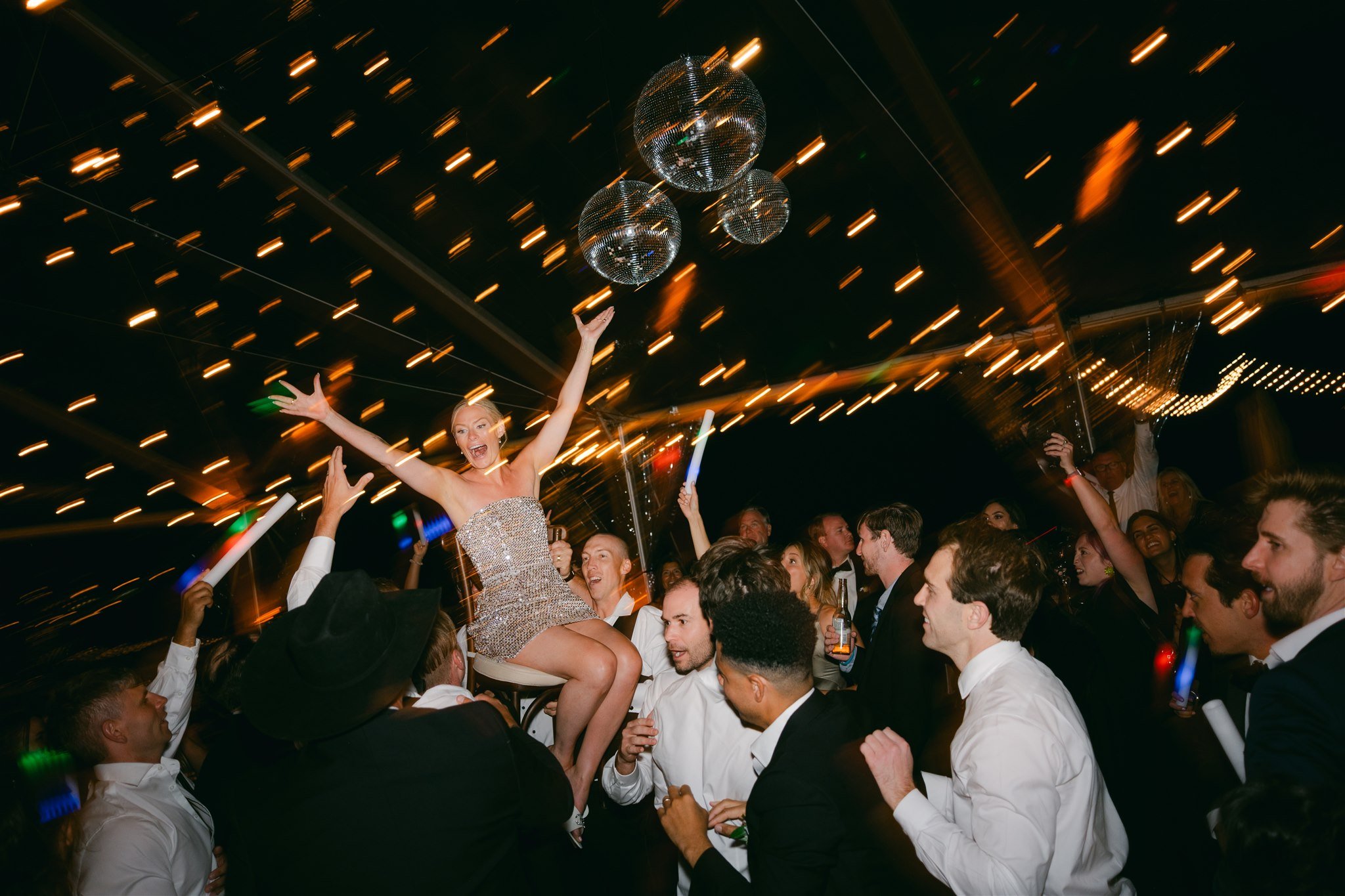 Bride in sequin reception dress being lifted on a chair by wedding guests on the dance floor under disco balls and string lights at Edgewood Tahoe Resort — Lake Tahoe wedding reception