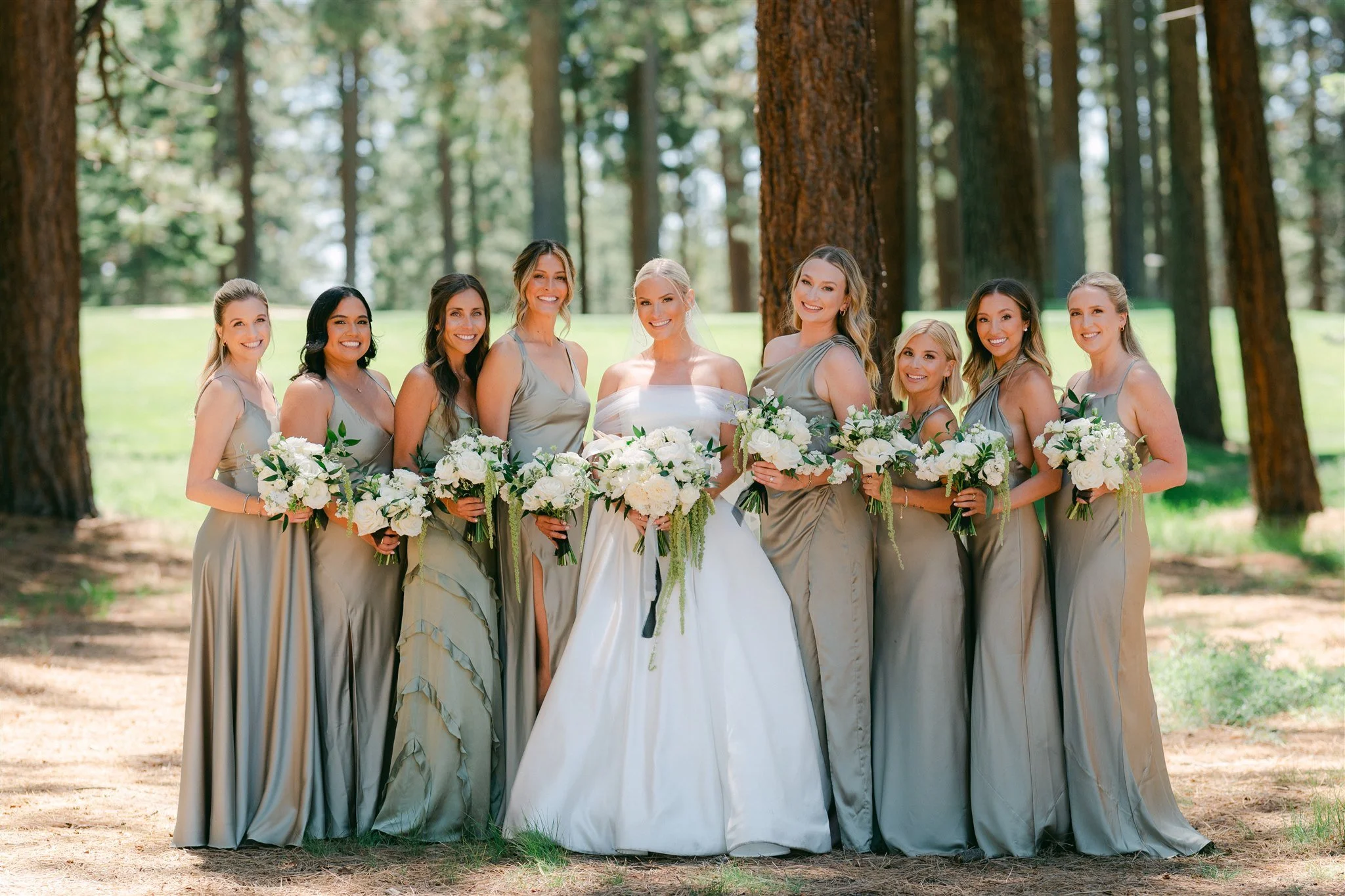 Bride in off-shoulder white ballgown surrounded by eight bridesmaids in sage taupe satin gowns holding white and green bouquets in a Tahoe pine forest — Edgewood Tahoe wedding