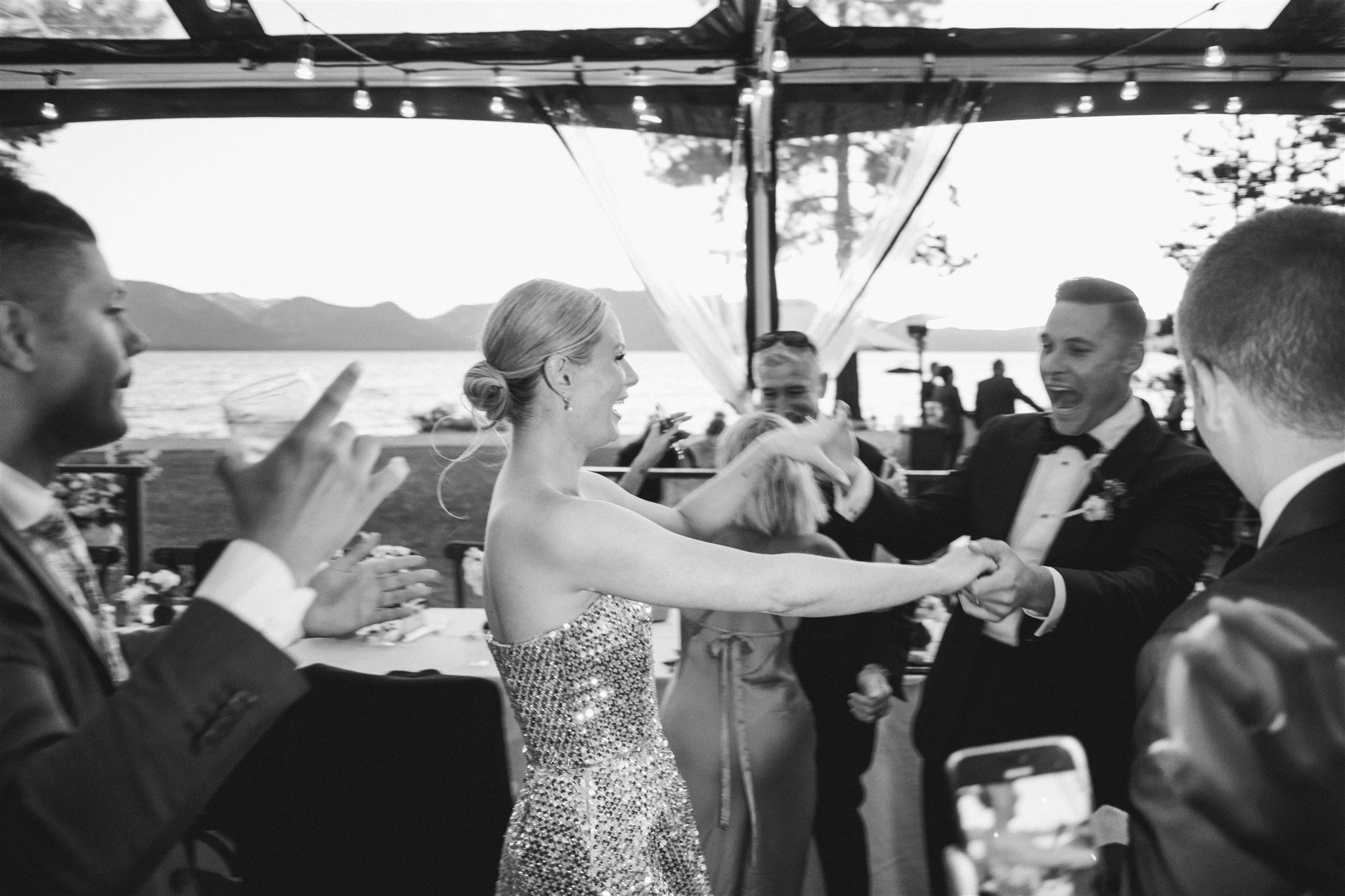 Black and white photo of bride in sparkly reception mini dress with arms raised dancing on the wedding dance floor under string lights at Edgewood Tahoe Resort