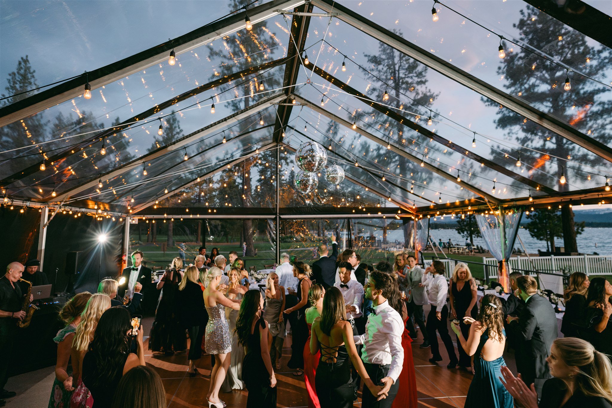 Full wedding dance floor at dusk under clear tent at Edgewood Tahoe Resort with live band, Edison string lights, cluster disco balls, and Lake Tahoe visible through tent walls — Sounds Elevated band