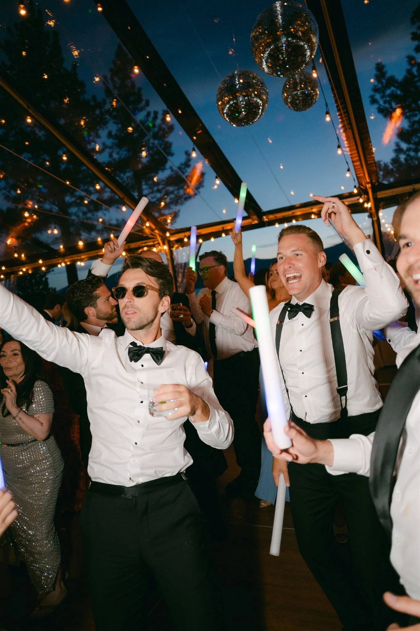 Wedding guests in tuxedos dancing with glow sticks under disco balls and string lights inside clear tent at Edgewood Tahoe Resort nighttime reception