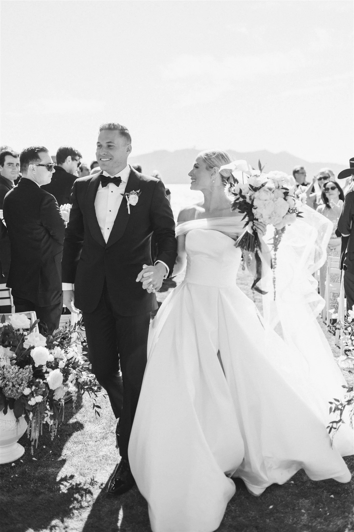Bride and groom sharing first kiss after outdoor Lake Tahoe wedding ceremony, photographed from behind with white ballgown train, guests standing and applauding, Lake Tahoe and pine trees in background