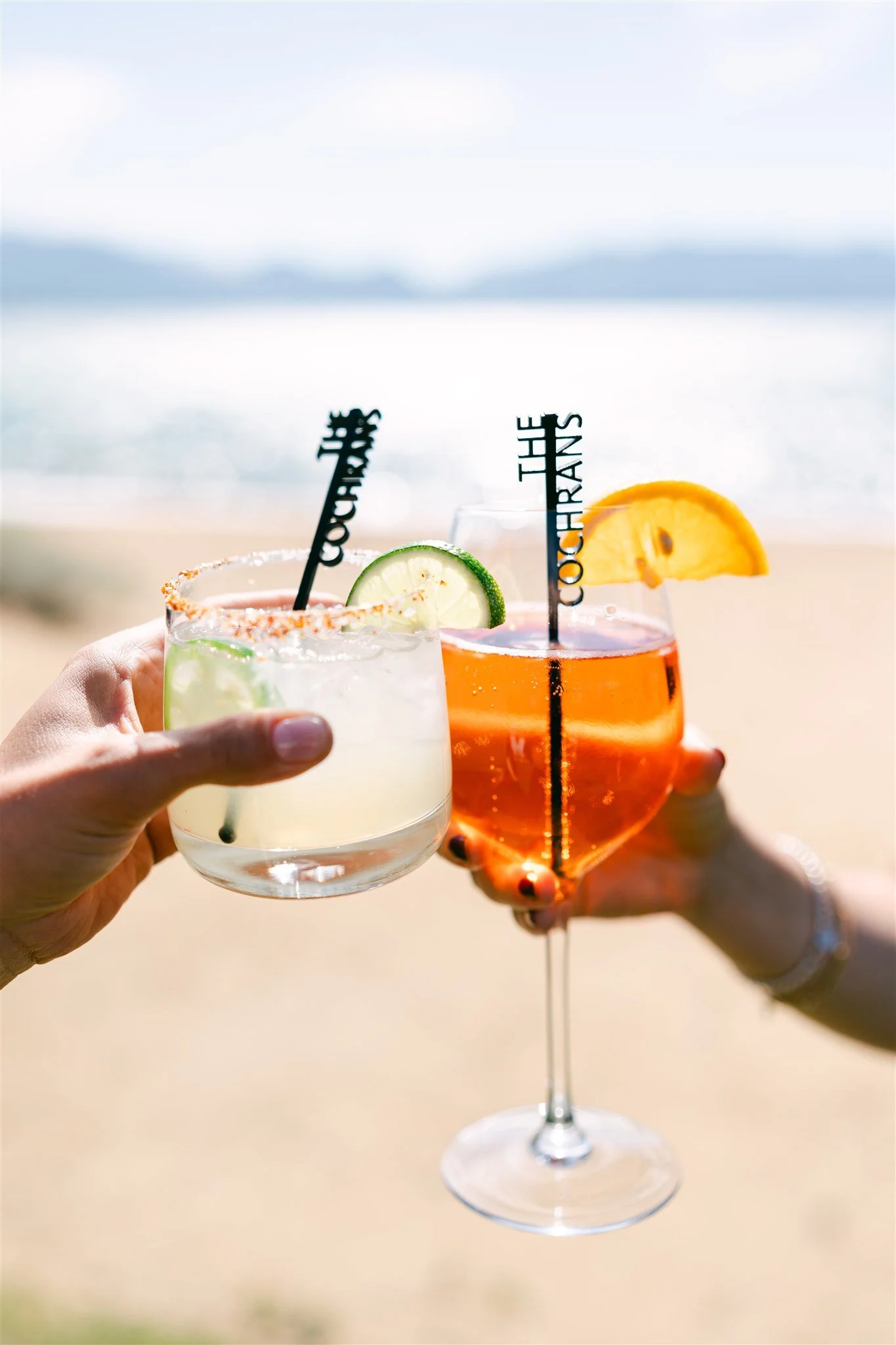 Two personalized wedding signature cocktails — a margarita and an Aperol spritz — being held up in a cheers on the Lake Tahoe beach with custom The Cochrans cocktail stirrers